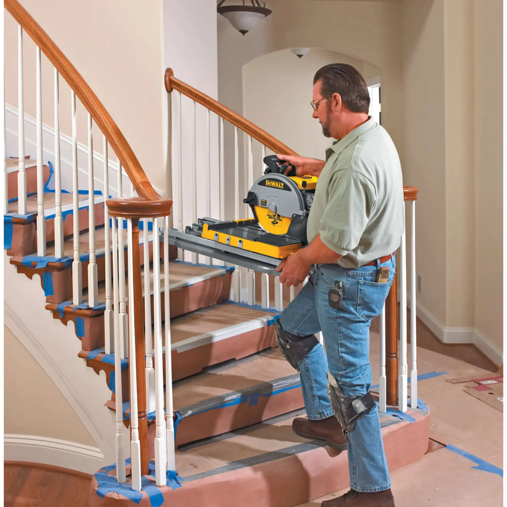 Una persona con rodilleras lleva una sierra para azulejos DEWALT D24000 por una escalera. Los escalones están cubiertos con papel protector y cinta azul. El entorno parece ser una obra en interiores durante una renovación.