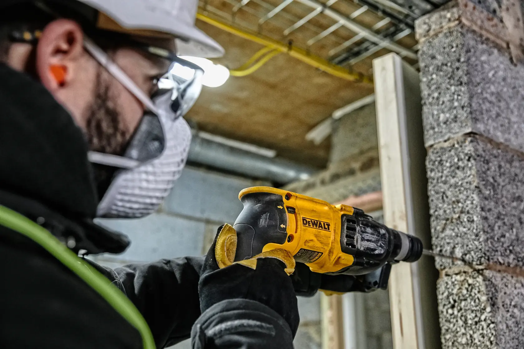 Un trabajador con equipo de seguridad utiliza un martillo perforador amarillo DEWALT (modelo D25133K) para perforar una pared de hormigón en una obra de construcción.