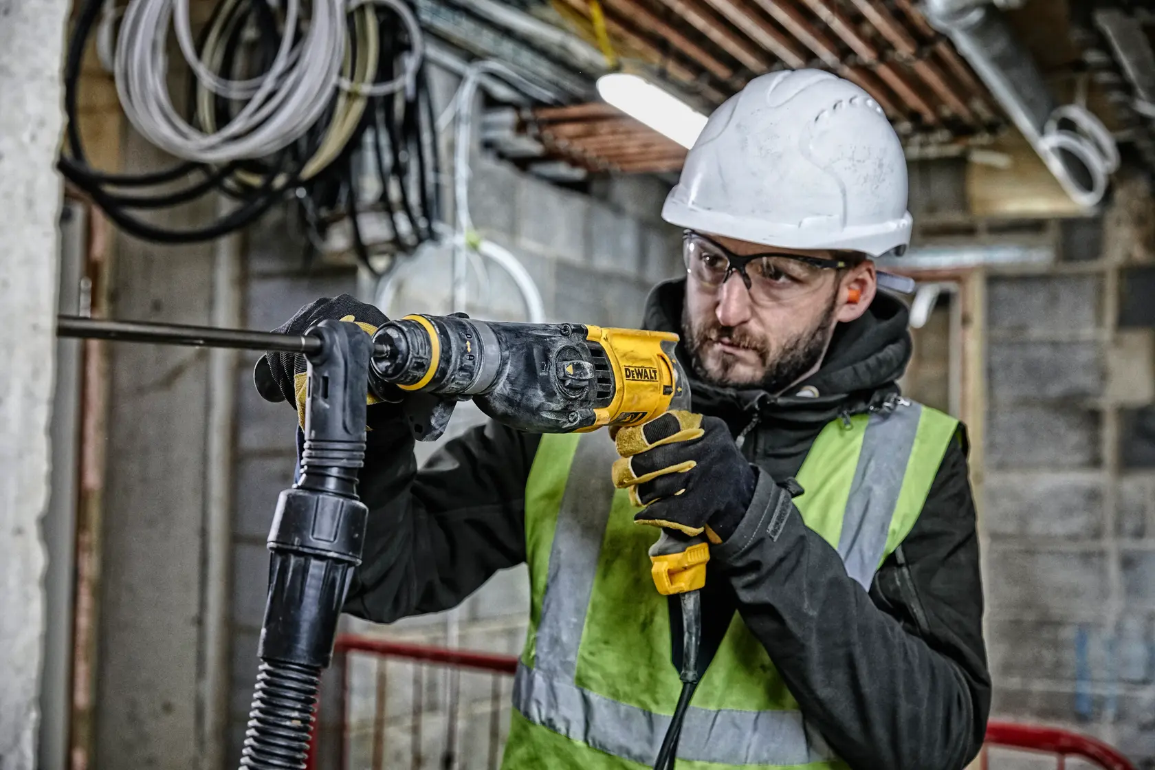 Una persona con casco de seguridad, guantes y chaleco reflectante utiliza un martillo rotativo DEWALT D25134K en una obra de construcción. Al fondo se observan cables y tuberías expuestos en un edificio sin terminar.