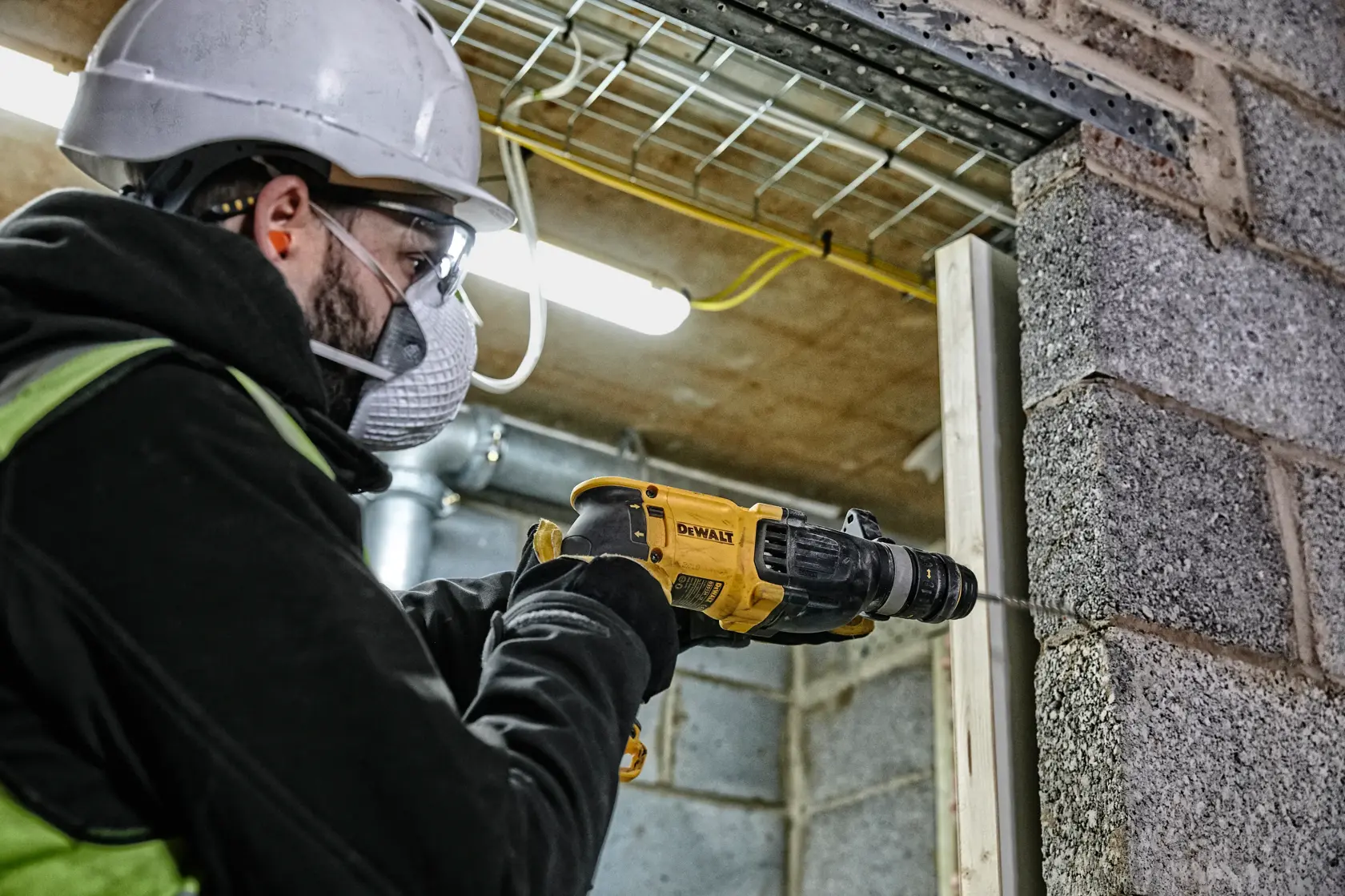 Un trabajador con casco de seguridad y guantes utiliza un martillo rotatorio DEWALT D25134K para perforar una pared de hormigón en una obra de construcción.