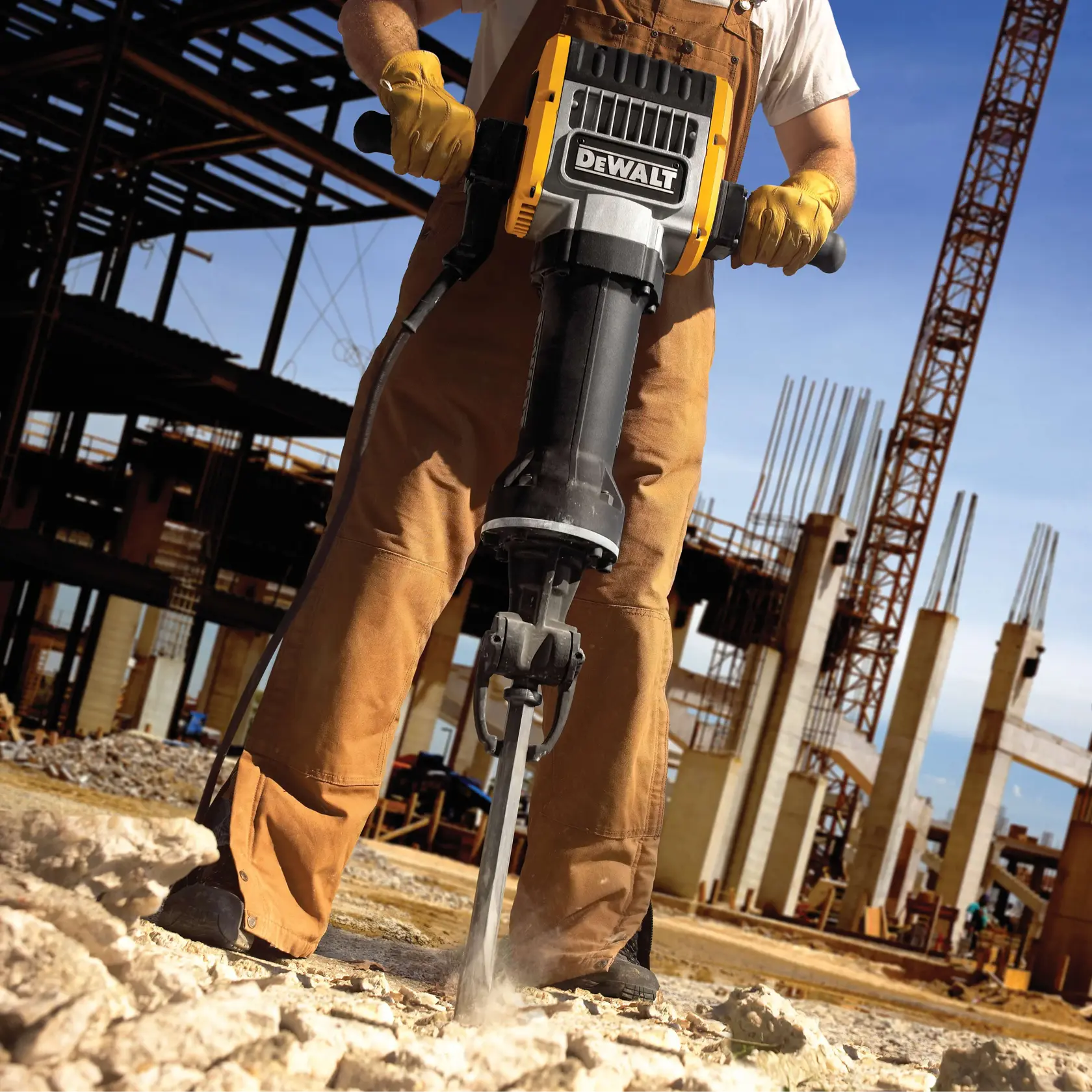 Una persona con guantes de trabajo y un overol opera un martillo neumático DEWALT, rompiendo concreto en un sitio de construcción, con estructuras de acero y columnas visibles al fondo.