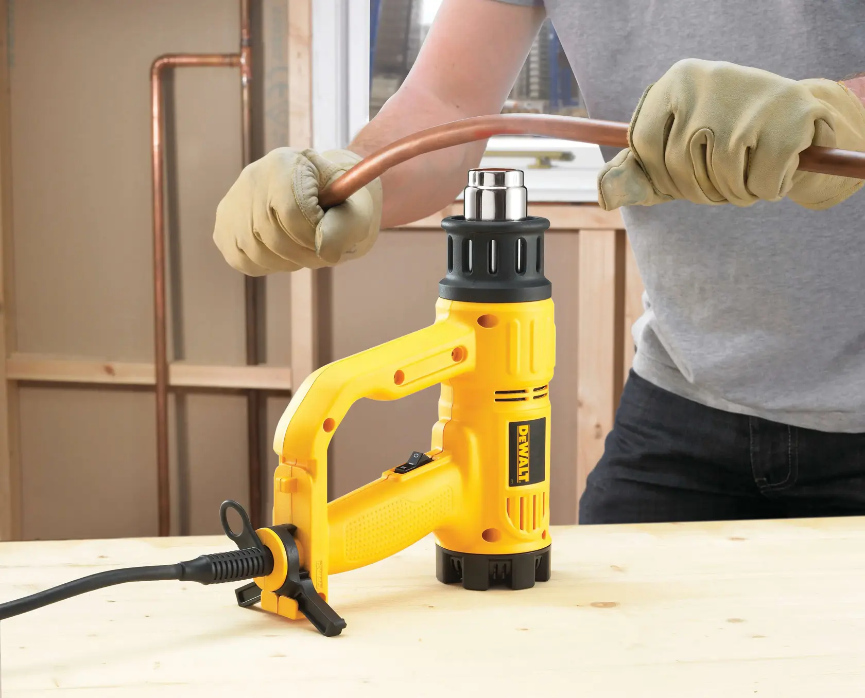 Una persona con guantes utiliza una pistola de calor DEWALT para doblar una tubería de cobre sobre una superficie de trabajo de madera.