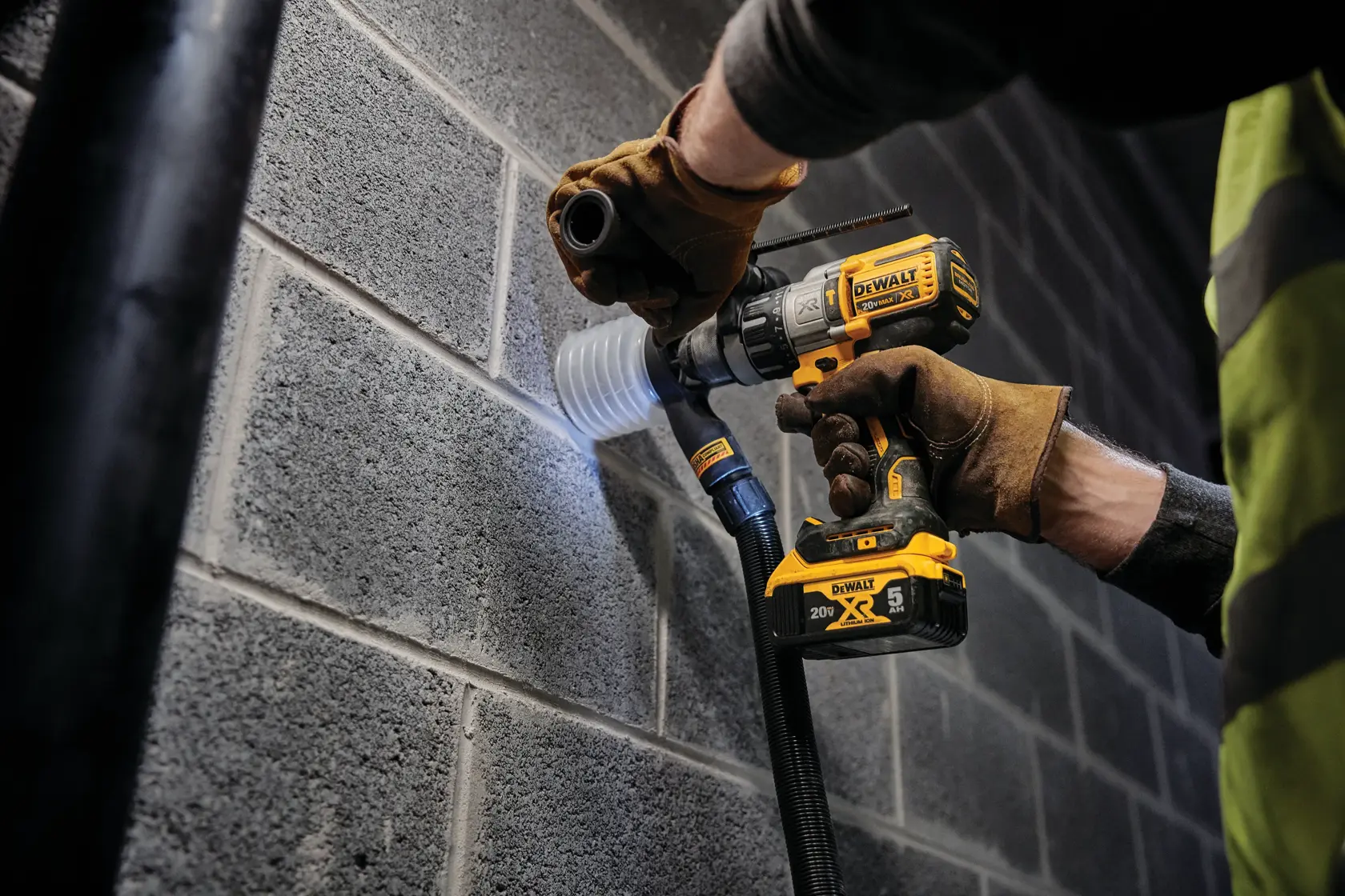 Una persona con guantes está usando un taladro inalámbrico DEWALT con una broca de corona para perforar una pared de bloques de cemento gris. Una manguera de extracción de polvo está conectada al taladro para recoger los residuos.