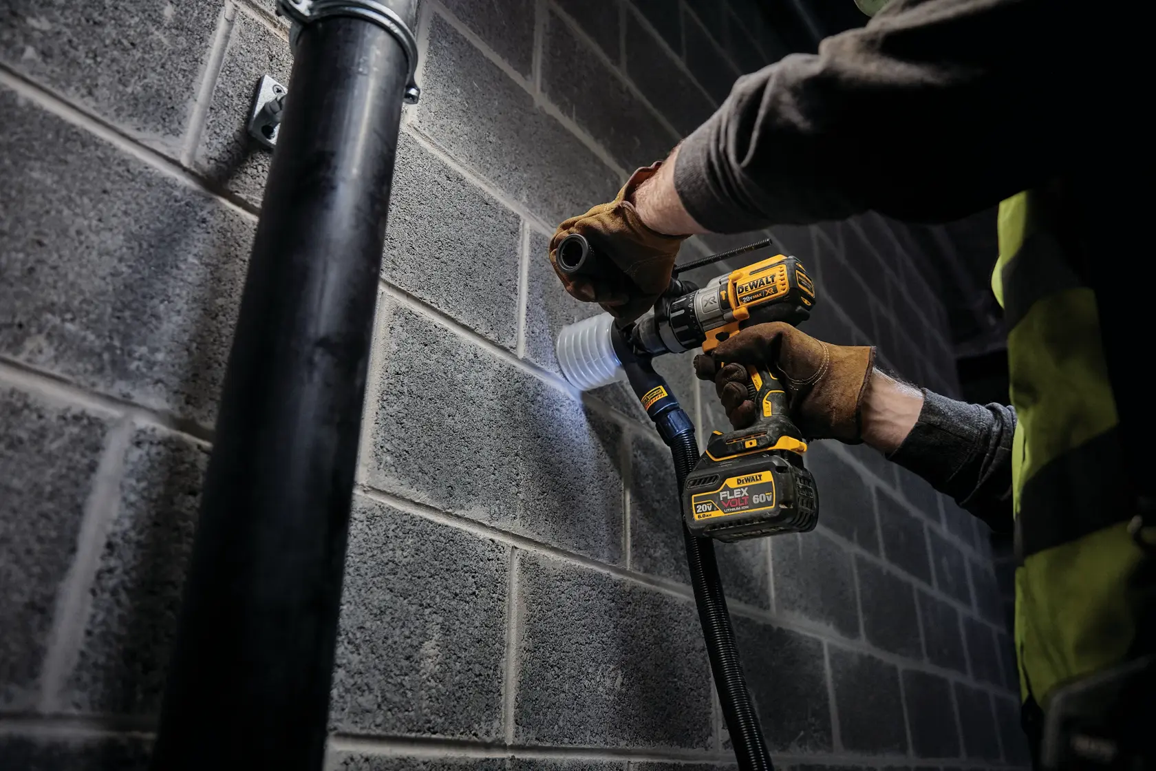 Una persona con guantes de trabajo utiliza un taladro inalámbrico DEWALT con accesorio de extracción de polvo para perforar una pared de bloques de cemento en una obra.