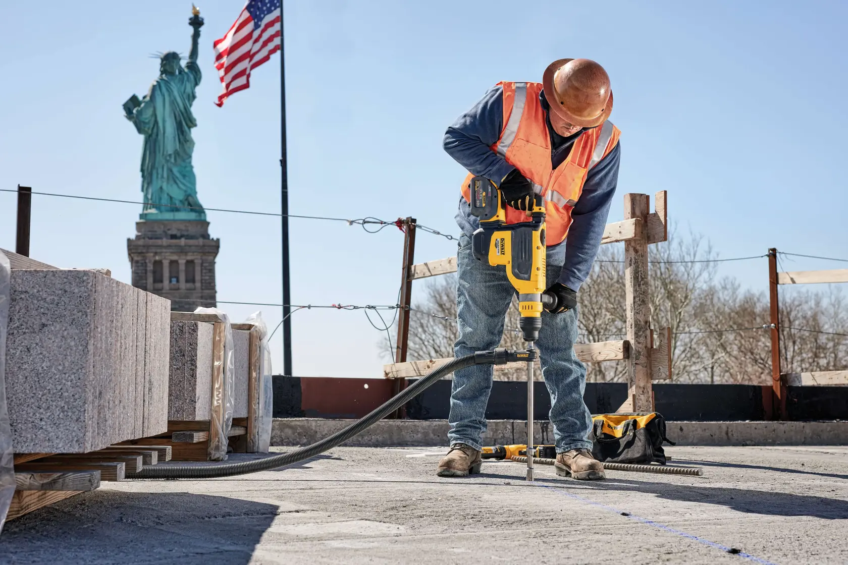 Un trabajador de la construcción con casco y chaleco de seguridad utiliza una herramienta eléctrica DEWALT para perforar concreto en una obra. Al fondo se observan la Estatua de la Libertad y una bandera estadounidense.