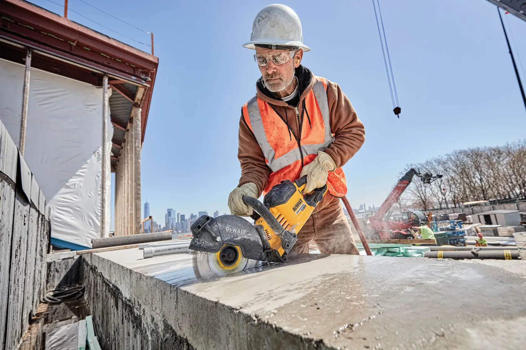 Un trabajador de la construcción con casco de seguridad y chaleco reflectante naranja utiliza una herramienta eléctrica DEWALT para cortar una losa de concreto en un sitio de construcción. La cara del trabajador está desenfocada y se ve polvo en el proceso de corte. De fondo se aprecian vigas estructurales, equipos y otros trabajadores.