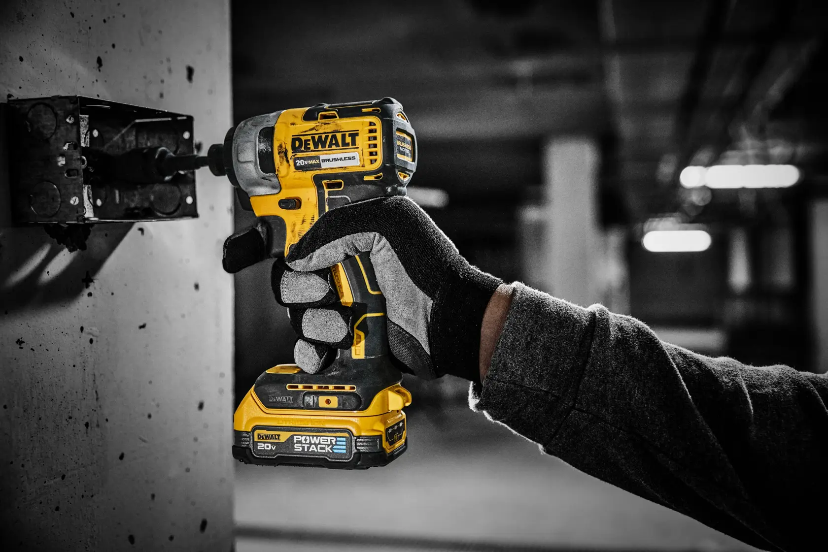 Una mano enguantada utiliza un atornillador de impacto inalámbrico DEWALT 20V amarillo y negro con batería POWERSTACK, atornillando un tornillo en una caja eléctrica de metal sobre una pared en un entorno de construcción o industrial.