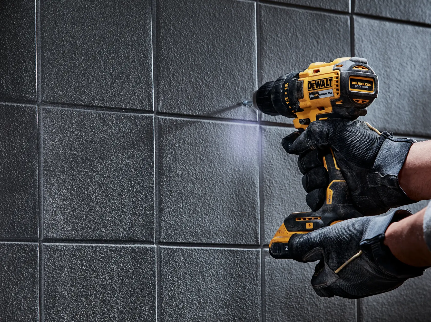 Una persona con guantes protectores negros está usando un taladro inalámbrico amarillo DEWALT para perforar una pared de azulejos grises.