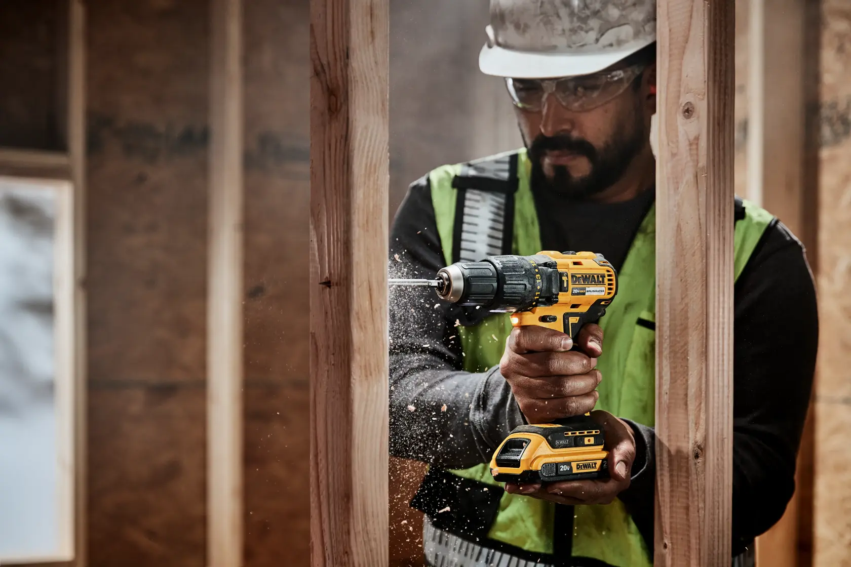 Una persona utiliza un taladro inalámbrico amarillo y negro DEWALT para perforar una tabla de madera en una obra de construcción. La persona lleva un chaleco de seguridad y un casco, y se puede ver aserrín en el aire.