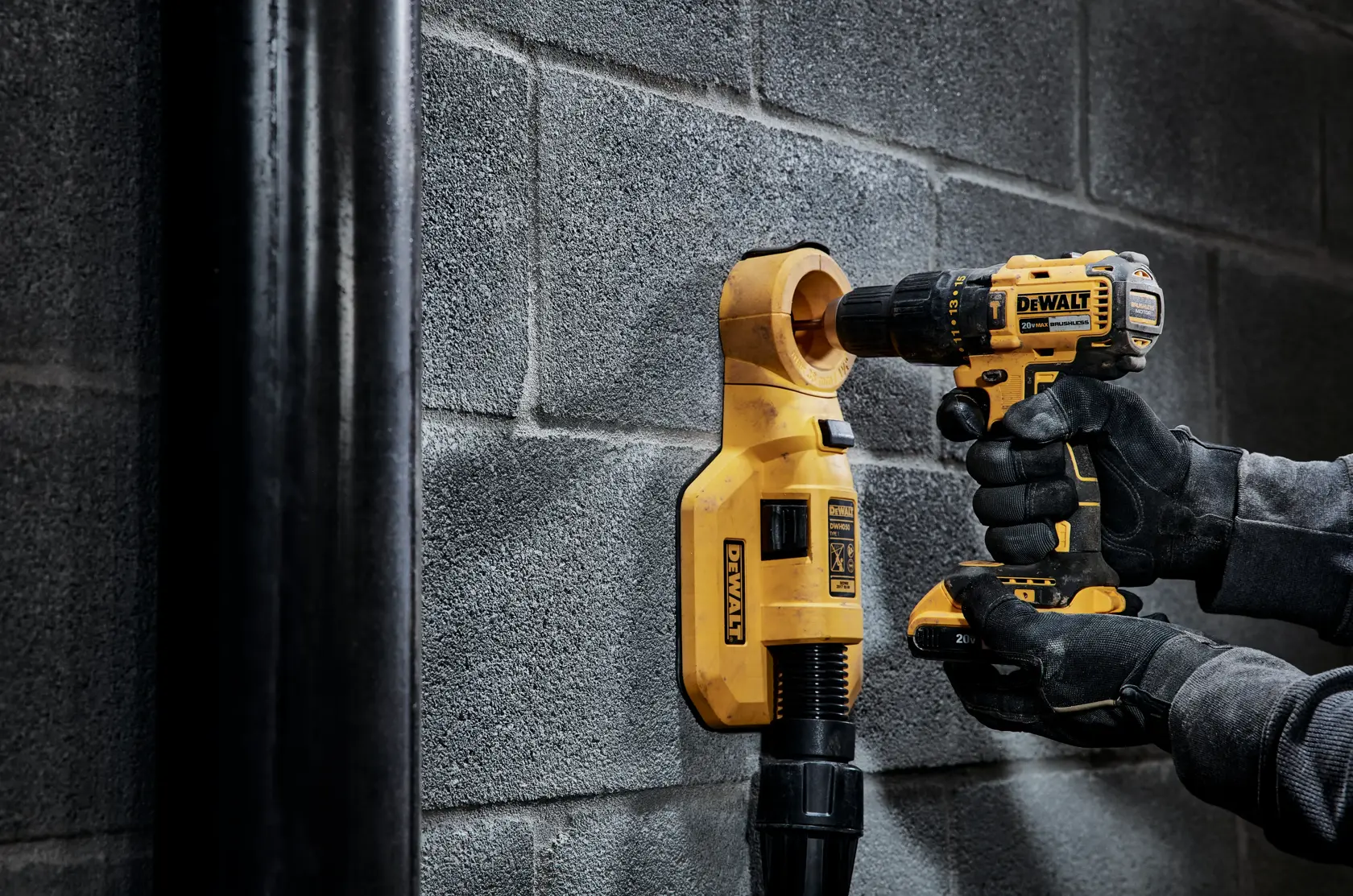Una persona con guantes negros está usando un taladro inalámbrico DEWALT para perforar una pared de bloques de concreto, con un accesorio de extracción de polvo amarillo DEWALT conectado debajo del taladro.