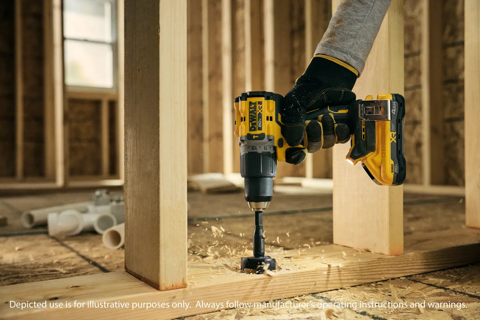 Una persona con guantes está usando un taladro inalámbrico amarillo y negro DEWALT para perforar un agujero en una viga de madera dentro de un edificio en construcción. Se ven virutas de madera alrededor de la broca. Hay tubos de PVC en el fondo.