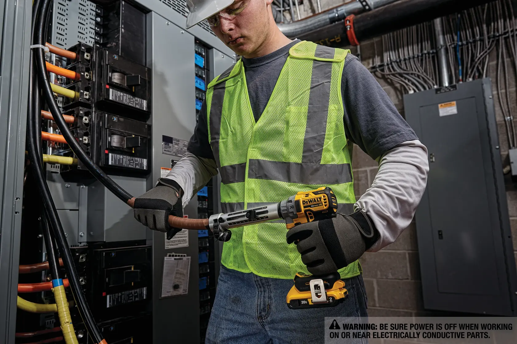 Una persona con chaleco de seguridad reflectante y guantes está utilizando una herramienta inalámbrica DEWALT para trabajar en cables eléctricos gruesos dentro de un panel eléctrico o caja de interruptores. El entorno parece ser una instalación eléctrica industrial o comercial con señales de advertencia visibles.