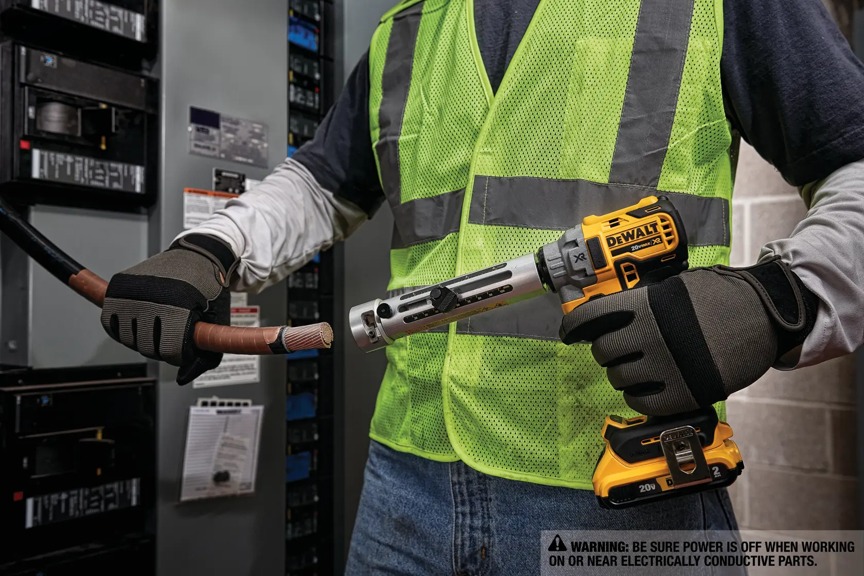 Una persona con guantes de seguridad y chaleco reflectante utiliza una herramienta eléctrica DEWALT para quitar el aislamiento de un cable eléctrico grueso frente a un panel eléctrico.