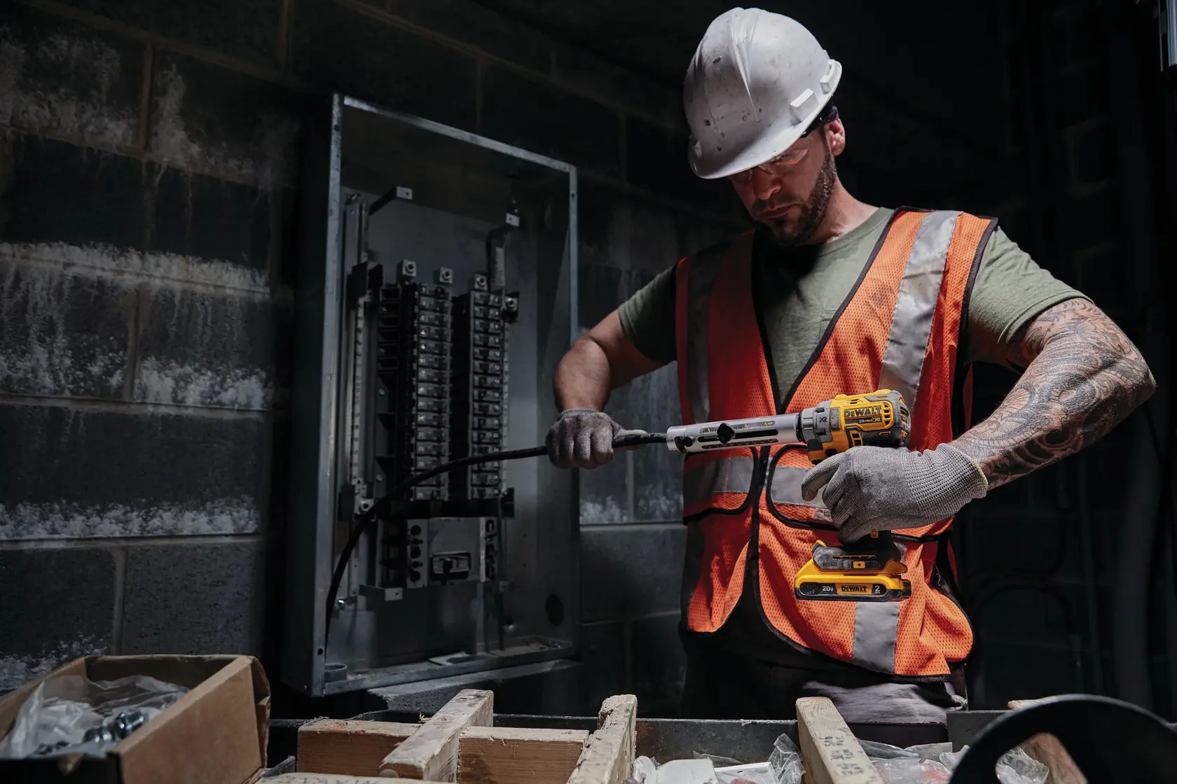 Un trabajador con casco y chaleco de seguridad naranja utiliza una herramienta inalámbrica DEWALT para trabajar en cableado eléctrico cerca de un panel de interruptores en un entorno industrial.