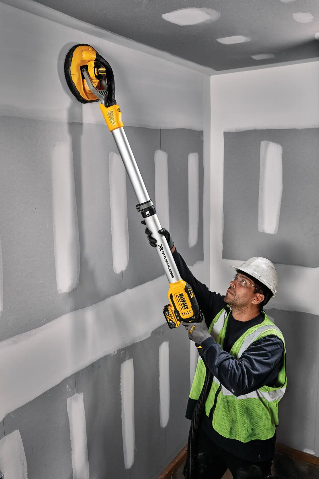 Un trabajador con chaleco de seguridad y casco usa una lijadora inalámbrica para paneles de yeso DEWALT DCE800P2 para lijar una pared y techo en una sala en construcción.