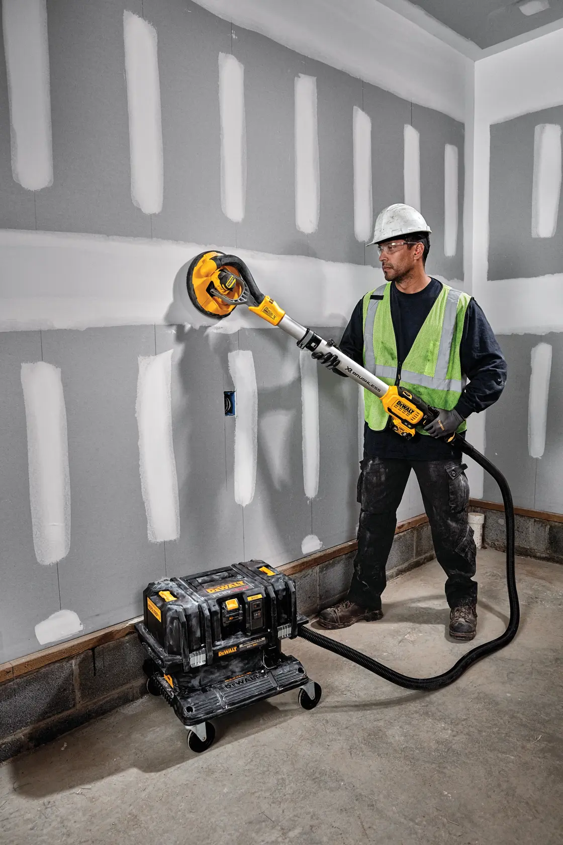 Un trabajador con chaleco de seguridad y casco utiliza una lijadora para paneles de yeso DEWALT con accesorio de aspiración, lijando una pared en un entorno de construcción. El equipo de aspiración está conectado a la lijadora y colocado en el suelo.