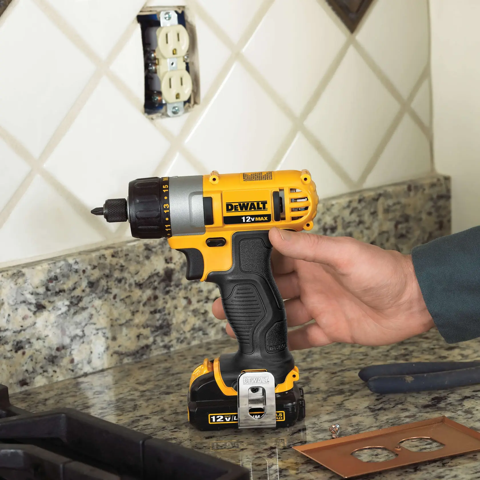 Una persona sostiene un taladro inalámbrico DeWalt 12V MAX amarillo y negro sobre una encimera de granito en la cocina, con una pared de baldosas. El taladro está cerca de una toma eléctrica en proceso de reparación y hay herramientas y cubiertas para tomas visibles cerca.