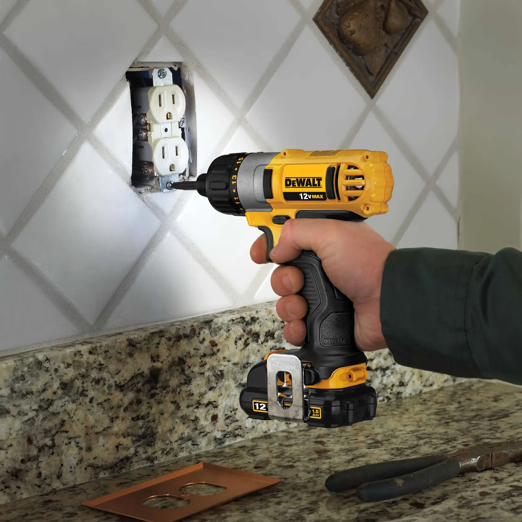 Una persona utiliza un taladro inalámbrico amarillo DEWALT 12V MAX para instalar o quitar tornillos de un enchufe eléctrico de pared, con el taladro colocado cerca de un salpicadero de azulejos y una encimera de granito.