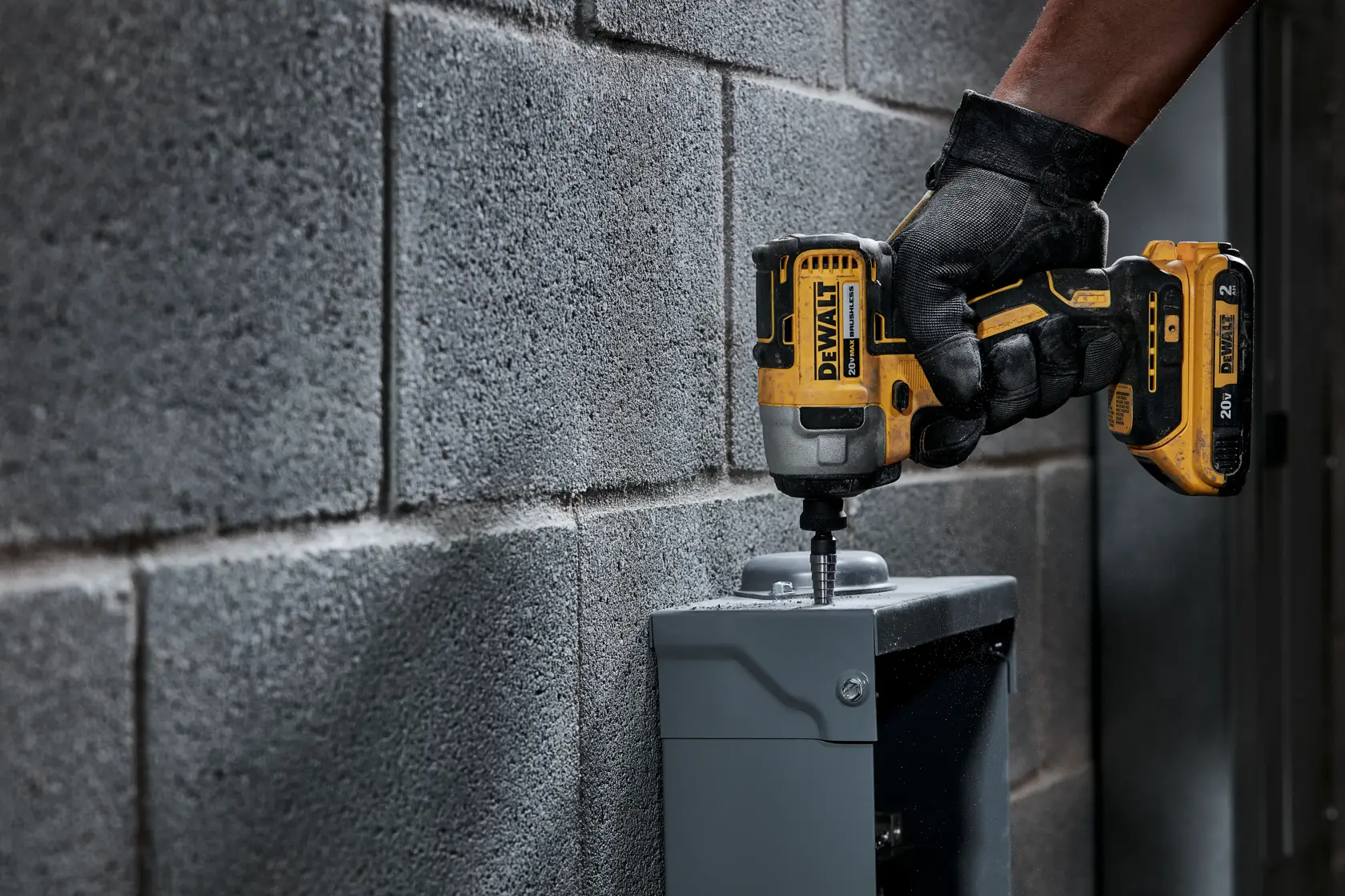 Una persona con guantes negros utiliza un atornillador de impacto inalámbrico amarillo DEWALT para fijar un tornillo en una caja metálica gris montada en una pared de bloques de hormigón.