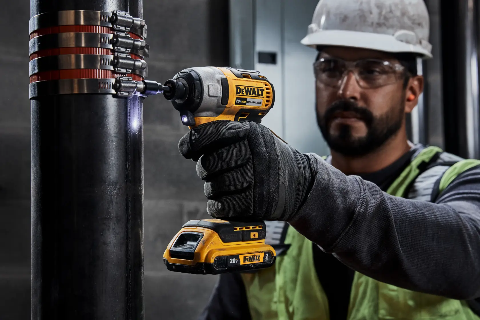 Una persona con casco y guantes de seguridad utiliza un atornillador de impacto inalámbrico DEWALT para ajustar abrazaderas en una tubería industrial grande. La herramienta DEWALT es de color amarillo y negro y se utiliza en un entorno de construcción o industrial.