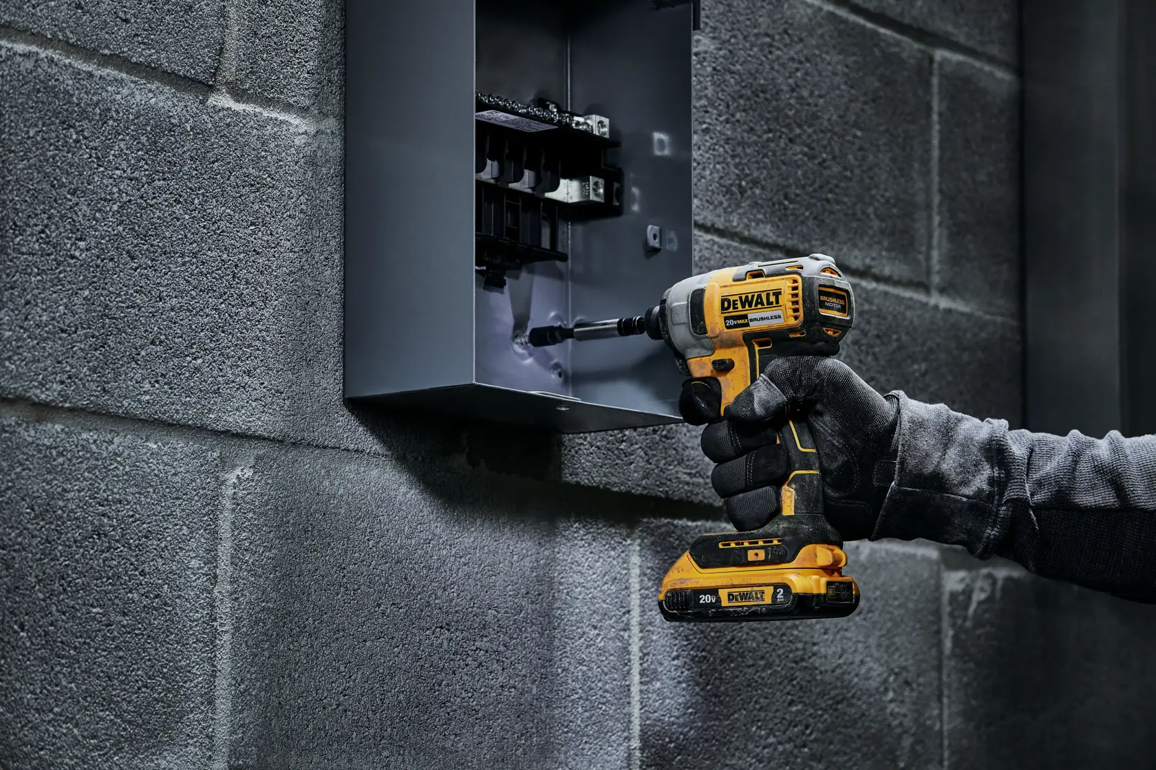 Una mano enguantada utiliza un taladro inalámbrico DEWALT para trabajar en un panel eléctrico montado en una pared de concreto gris.
