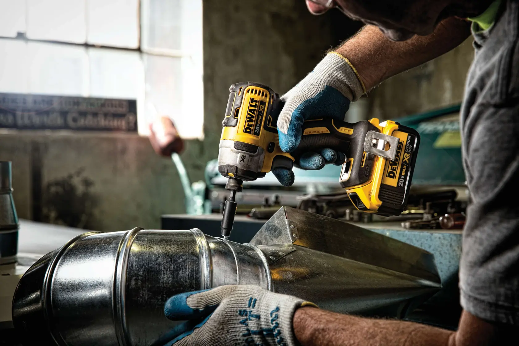Una persona con guantes de trabajo utiliza un destornillador de impacto inalámbrico DEWALT DCF887D2 para asegurar un tornillo en un conducto de metal en un taller industrial.