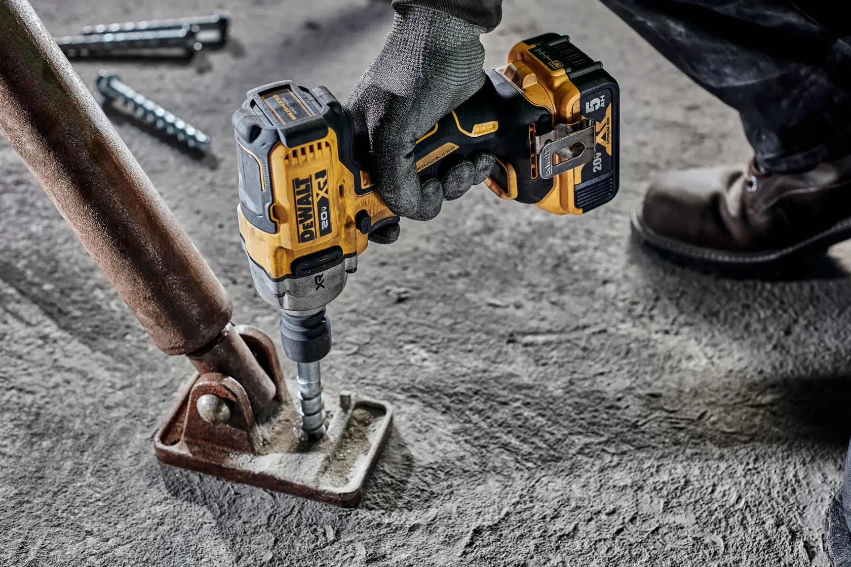 Una mano enguantada utiliza una herramienta eléctrica DEWALT con batería de 20V, fijando un tornillo en una base metálica sobre un suelo de cemento.