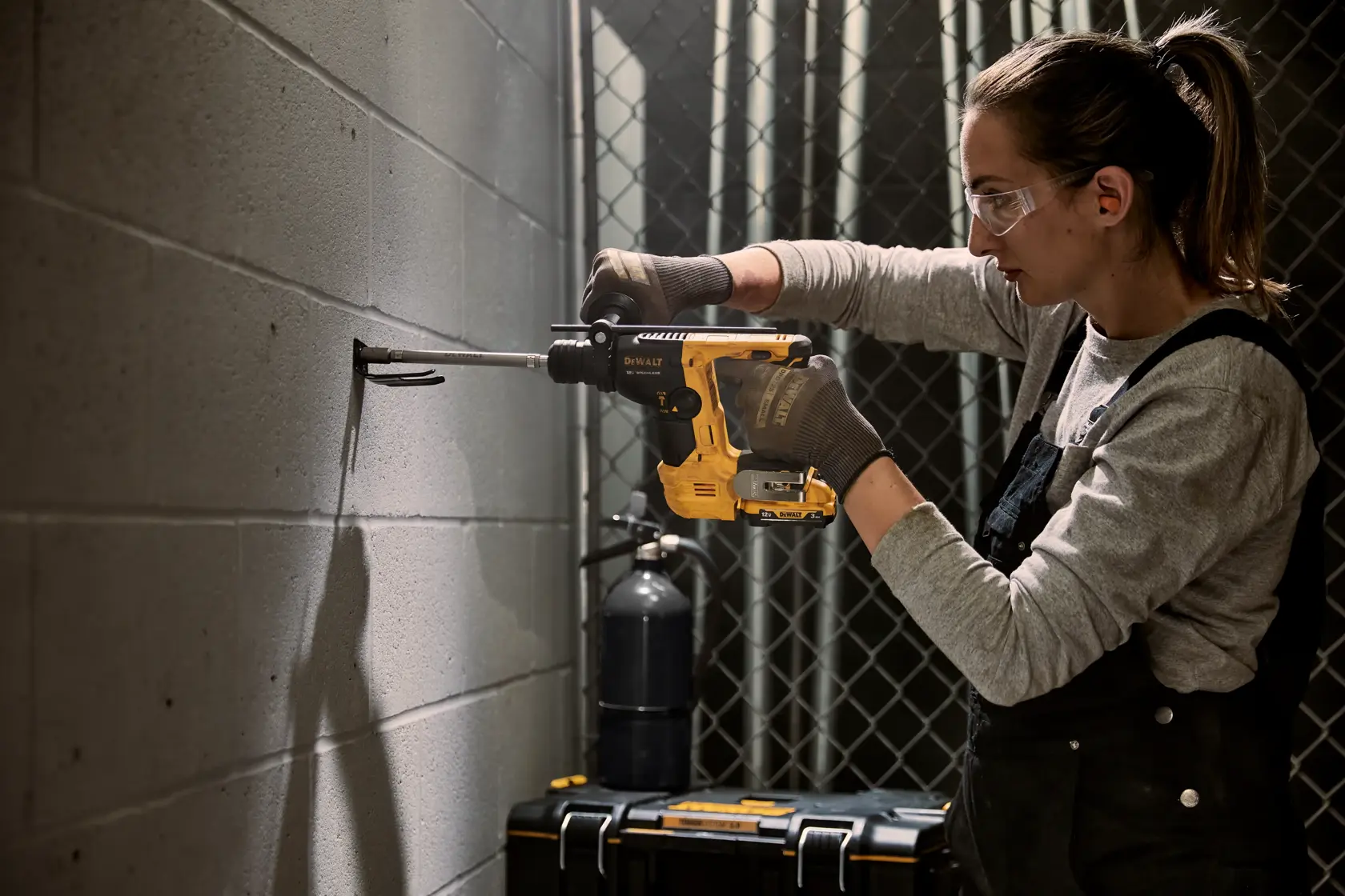 Una persona con guantes está usando un taladro eléctrico DEWALT para perforar una pared de concreto. La escena es en interiores con una caja de herramientas y otros equipos visibles en el fondo. El rostro de la persona está intencionadamente borroso.