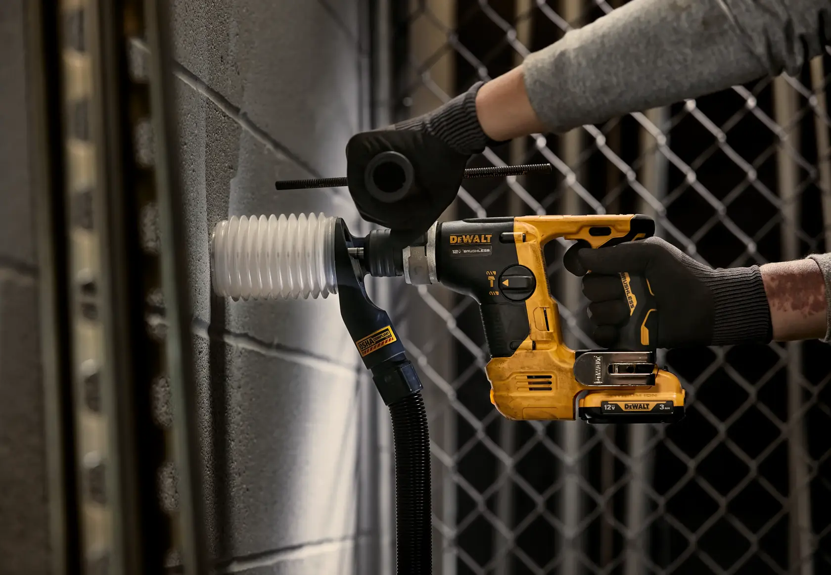 Una persona con guantes utiliza un taladro rotativo inalámbrico DEWALT con un accesorio de extracción de polvo para perforar una pared de concreto.