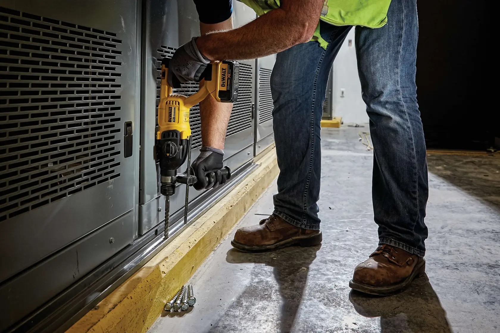 Una persona con jeans, botas de trabajo y guantes utiliza un martillo rotatorio inalámbrico DEWALT para perforar una superficie metálica cerca del suelo en un entorno industrial o de construcción.