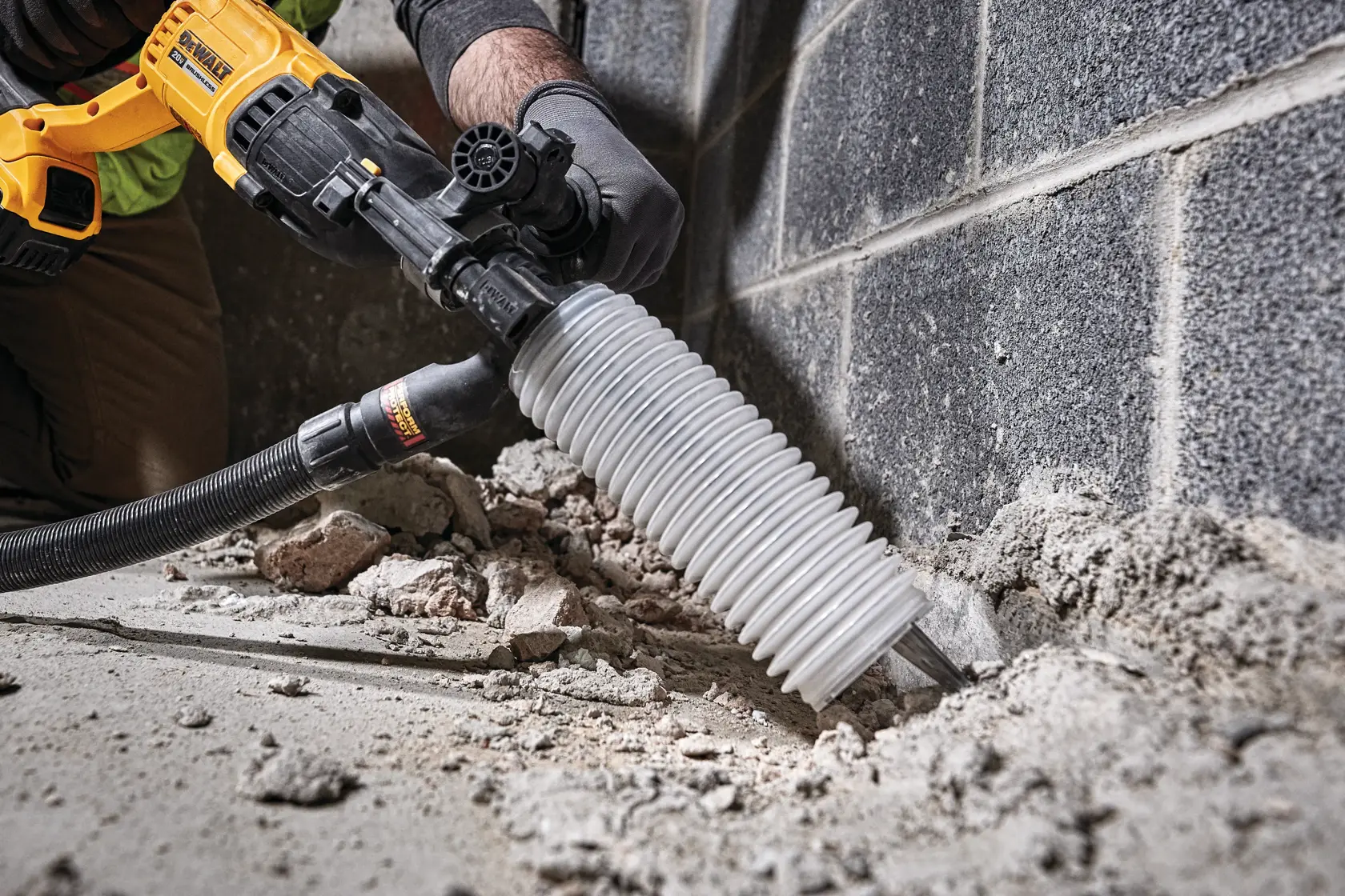 Una persona usa un martillo rotativo DEWALT con un accesorio para recolectar polvo para romper concreto cerca de una pared. La herramienta es de color amarillo y negro, y hay escombros visibles en el suelo.