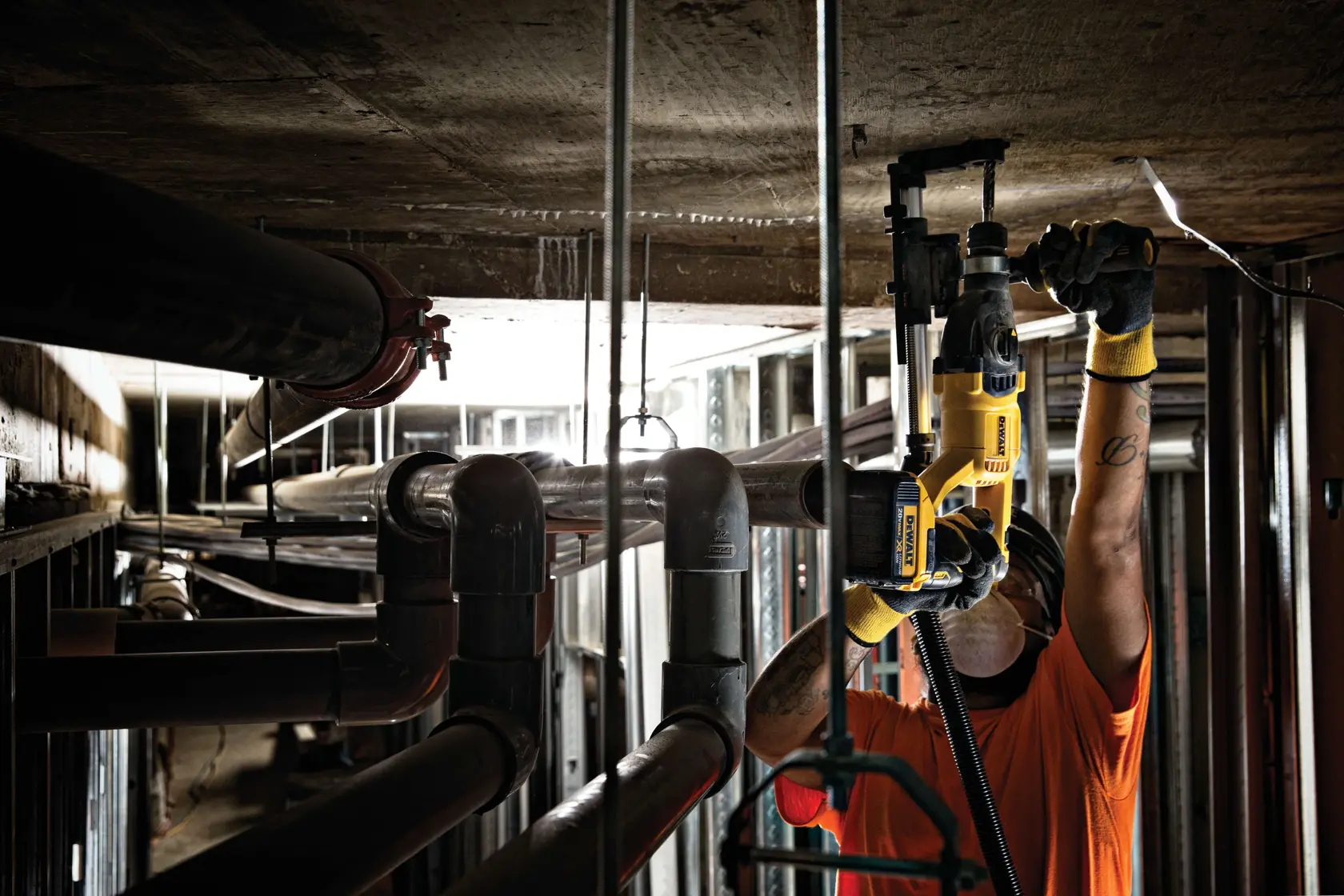 Una persona con casco de seguridad y guantes utiliza un taladro percutor amarillo DEWALT para trabajar en un techo de concreto en un entorno industrial con tuberías y estructuras metálicas visibles.