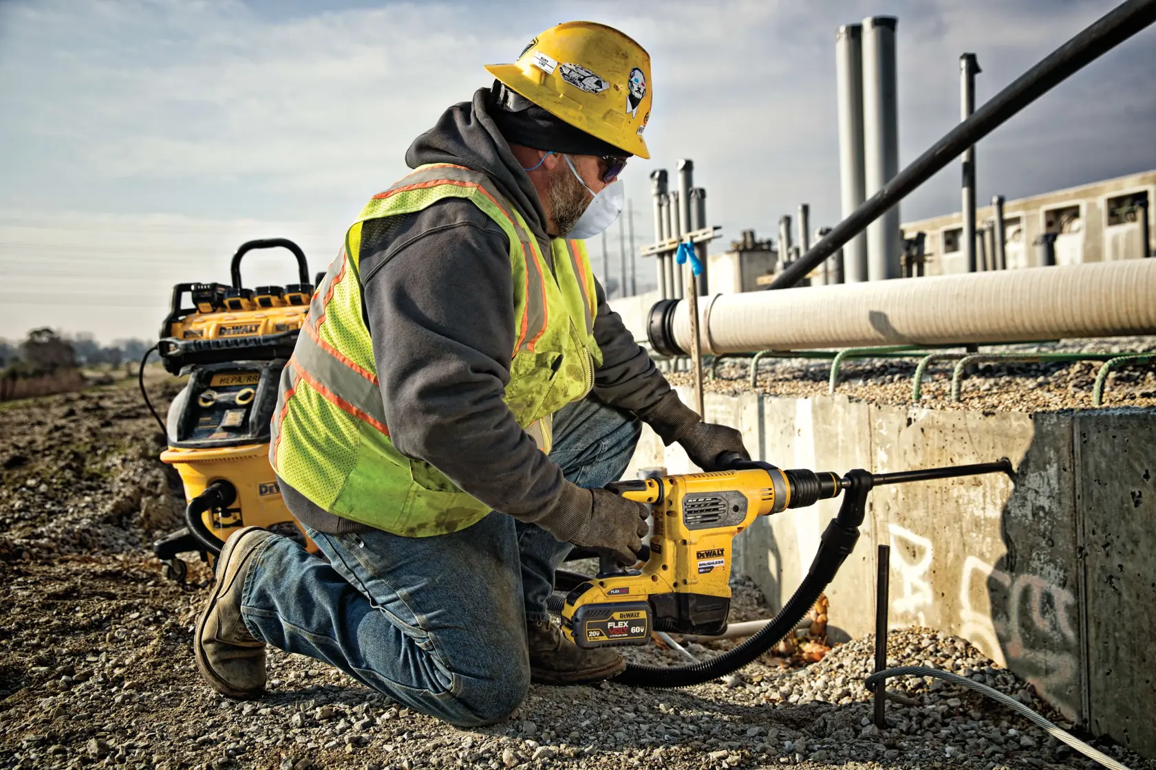 Un trabajador de la construcción con casco amarillo y chaleco de seguridad está arrodillado sobre grava mientras usa una herramienta eléctrica DEWALT para perforar una pared de concreto en un sitio de construcción al aire libre. El rostro del trabajador está difuminado por privacidad.