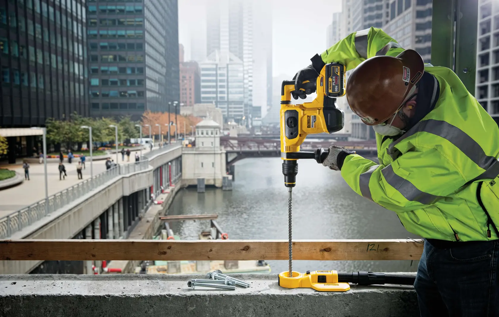 Una persona con chaqueta de alta visibilidad y casco está usando un taladro rotativo DEWALT DCH481B sobre una superficie de concreto en un sitio de construcción con vista a un río y un puente en la ciudad.