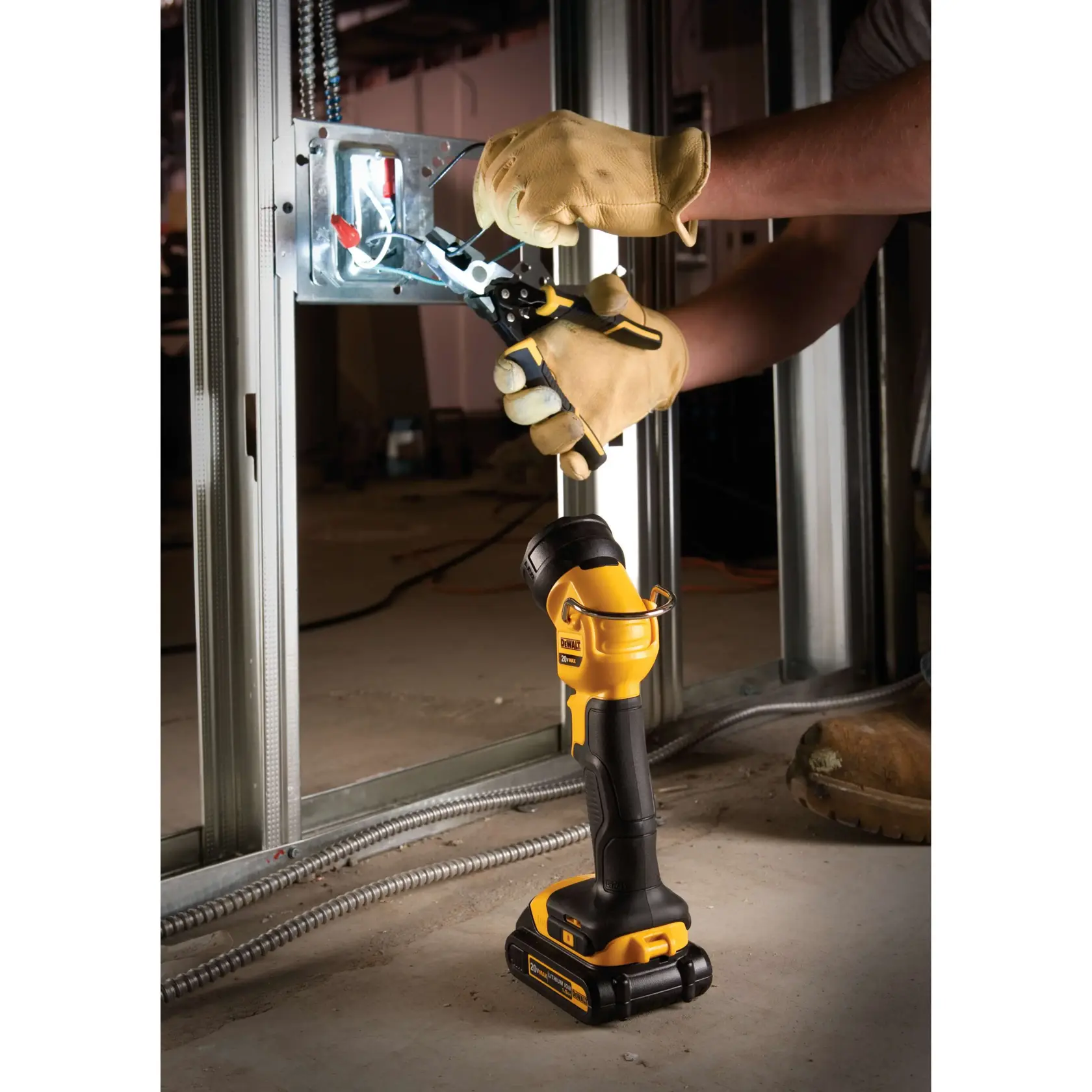 Una lámpara de trabajo LED portátil DEWALT DCL040 está colocada en el suelo, iluminando una caja eléctrica mientras una persona con guantes de trabajo utiliza unas pinzas para trabajar en el cableado dentro de un marco de pared metálico.