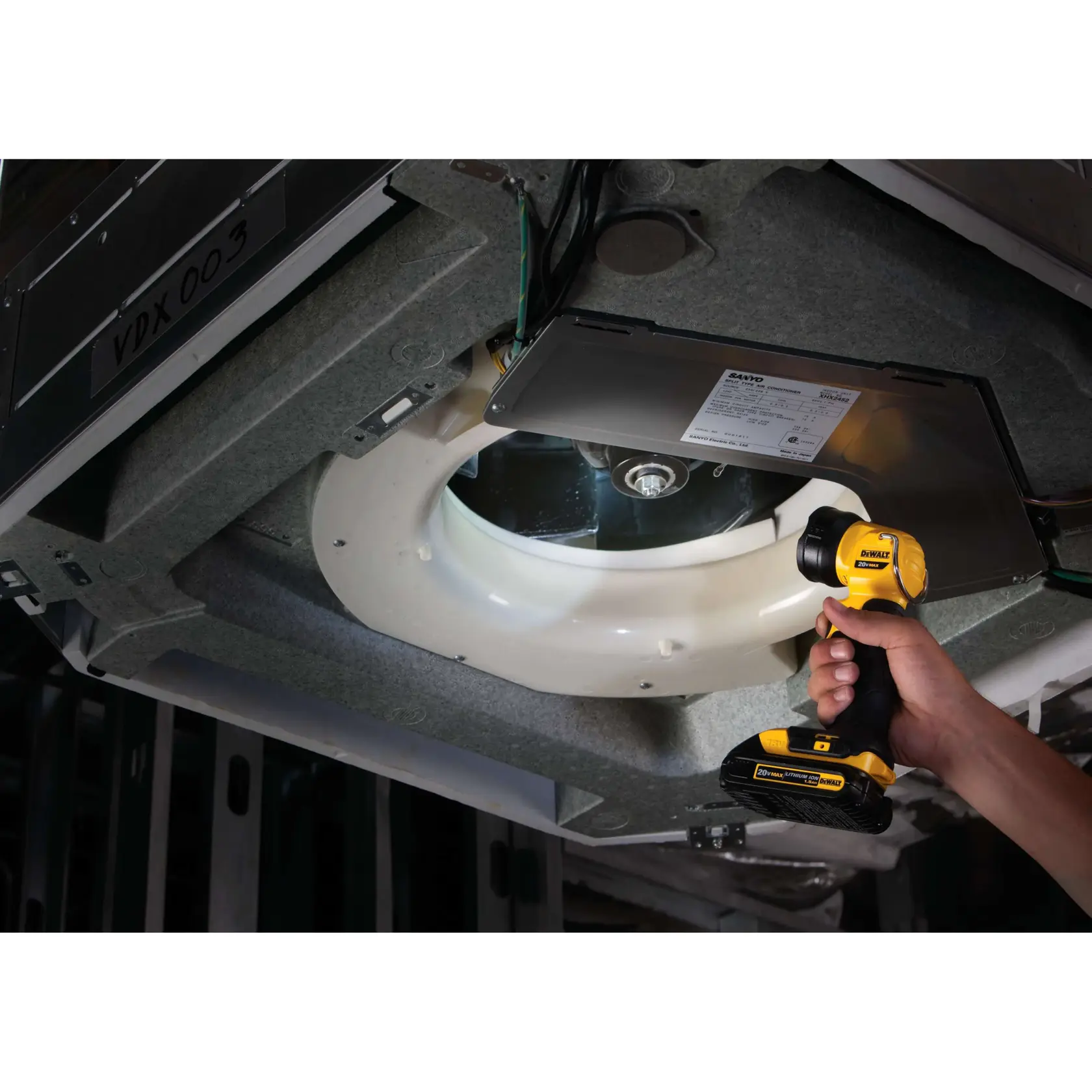 Una persona utiliza una lámpara LED portátil de trabajo DEWALT DCL040 para iluminar los componentes internos de una unidad de aire acondicionado o equipo mecánico similar durante el mantenimiento.