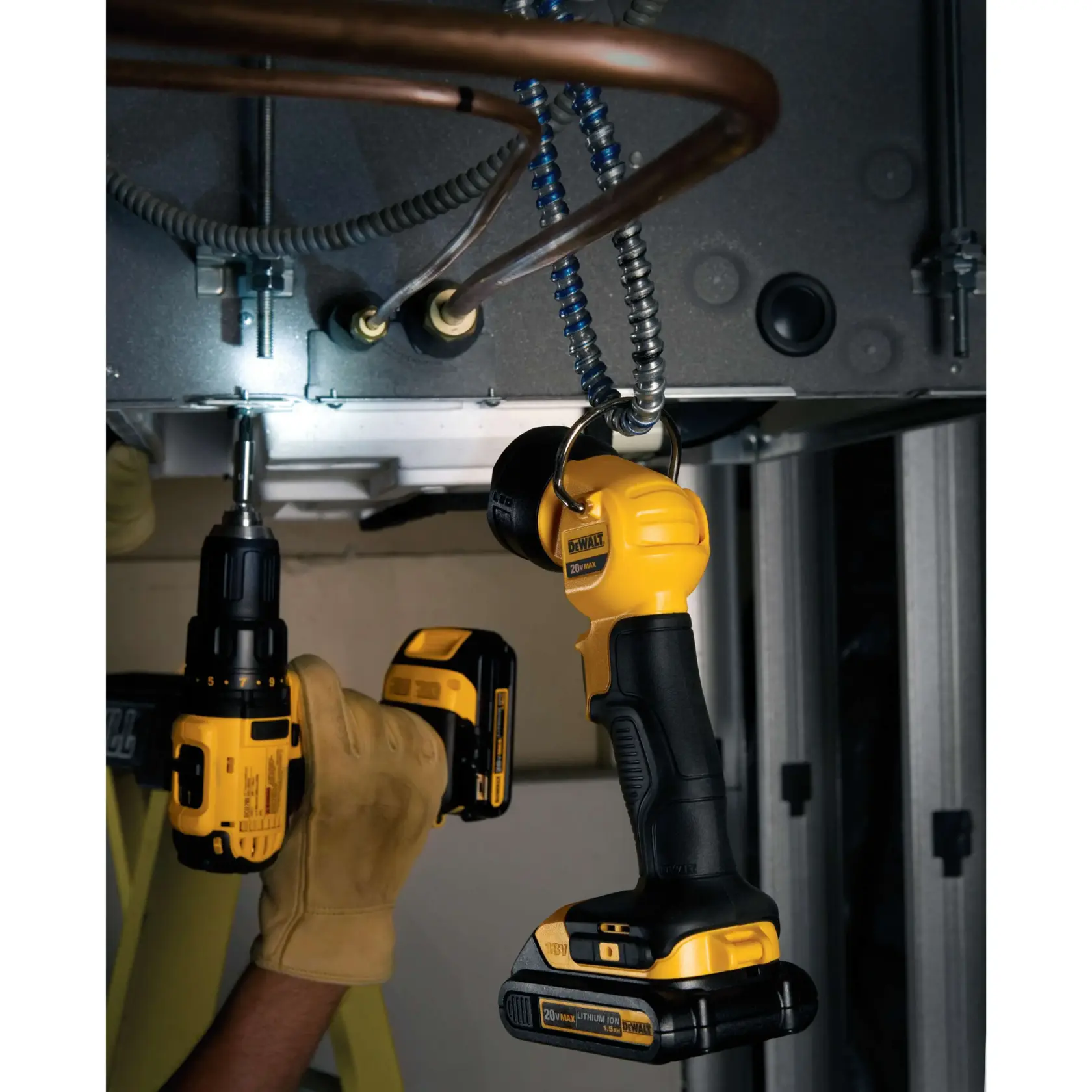 Una lámpara de trabajo LED inalámbrica DEWALT (modelo DCL040) cuelga de un accesorio metálico, iluminando el área de trabajo. Cerca, una persona con guantes de trabajo beige utiliza un taladro inalámbrico DEWALT para trabajar sobre equipos y tuberías metálicas.
