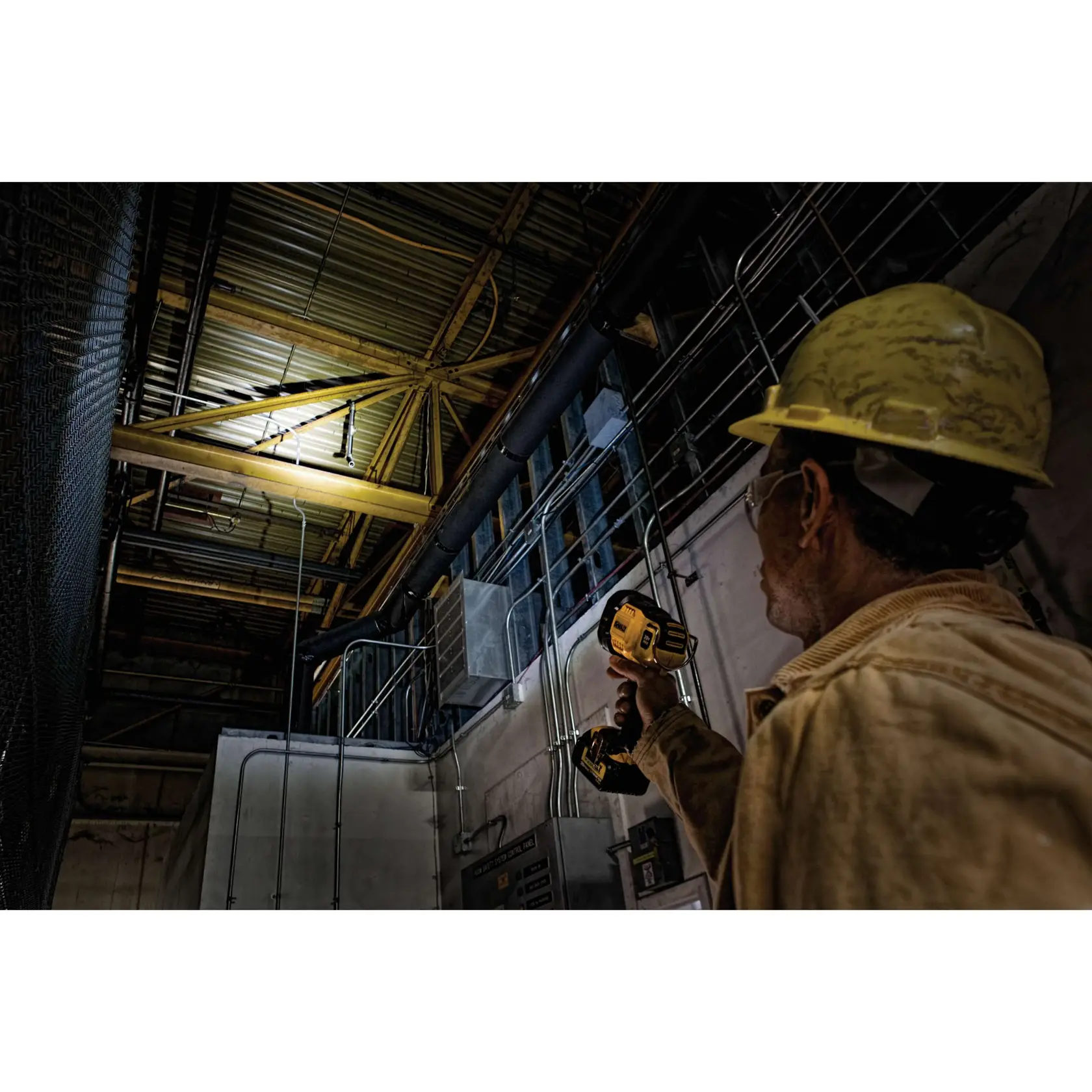 Un trabajador con casco y chaqueta amarillos sostiene una linterna DEWALT DCL043 y la dirige hacia el techo de un edificio industrial, iluminando tuberías y vigas metálicas.