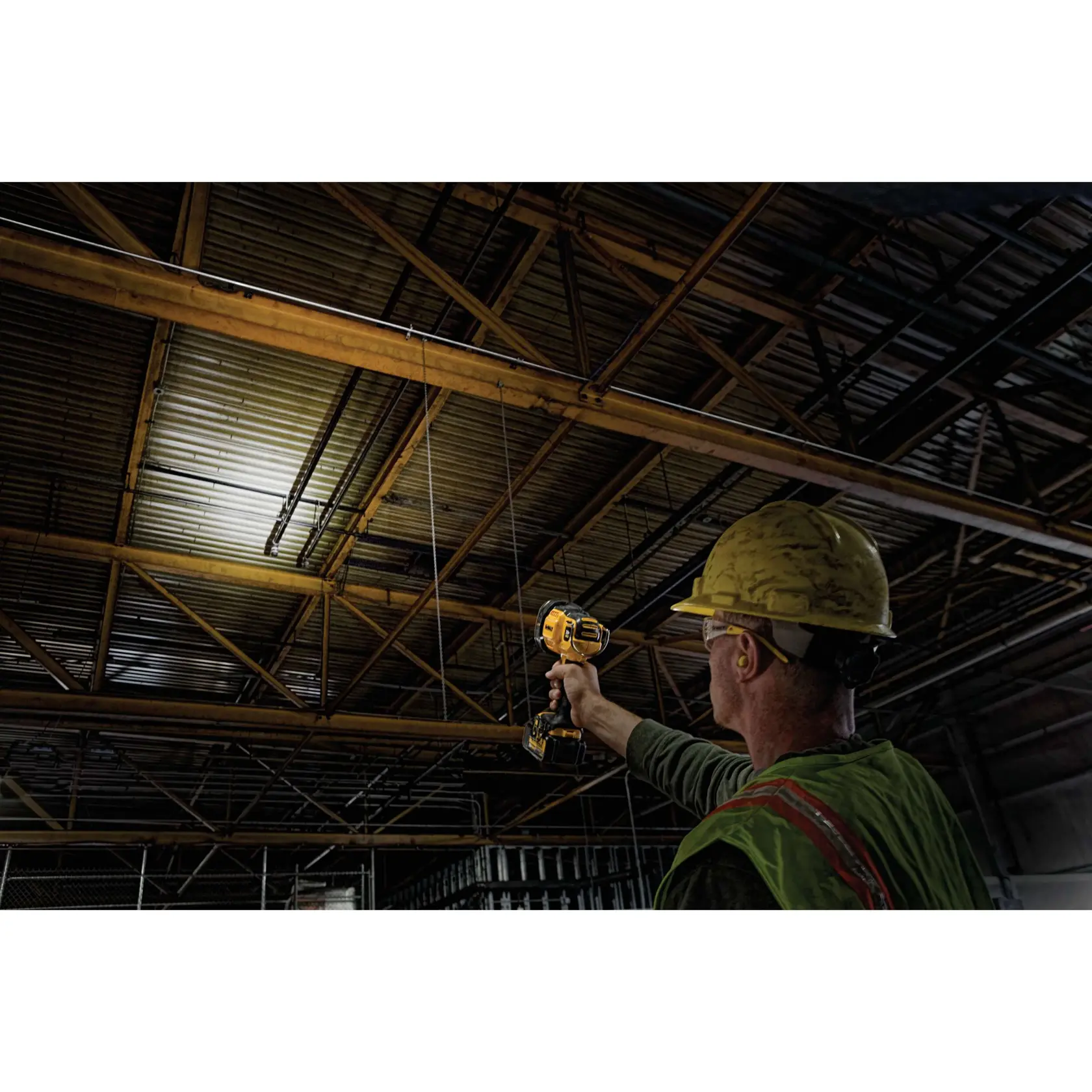 Un trabajador con casco de seguridad amarillo y chaleco utiliza un foco portátil DEWALT para iluminar la estructura del techo de un edificio industrial.