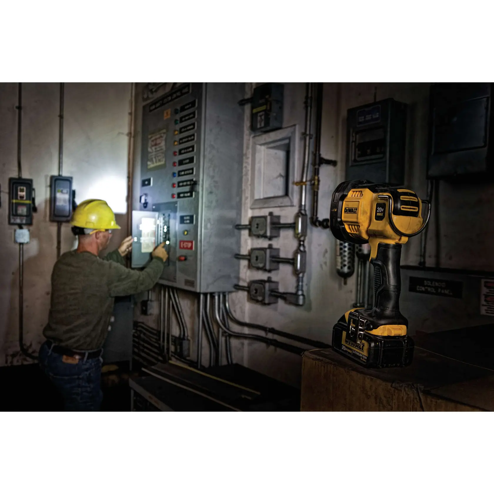 Una lámpara de trabajo portátil DEWALT modelo DCL043 está colocada sobre una superficie en un entorno industrial. En el fondo, un trabajador con casco amarillo opera un panel de control eléctrico. El área parece estar débilmente iluminada, con la lámpara proporcionando luz.