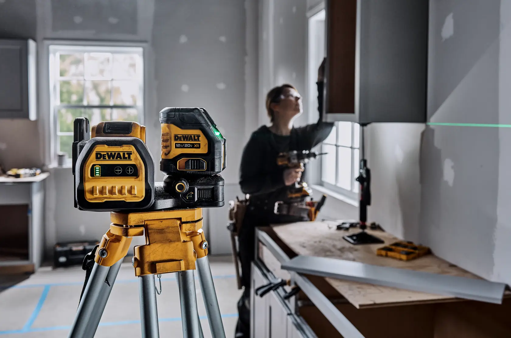 Dispositivo de nivel láser DEWALT montado en un trípode en primer plano, proyectando una línea láser verde sobre una pared en una habitación en construcción. Una persona trabaja en el fondo cerca de la ventana, instalando o ajustando gabinetes.