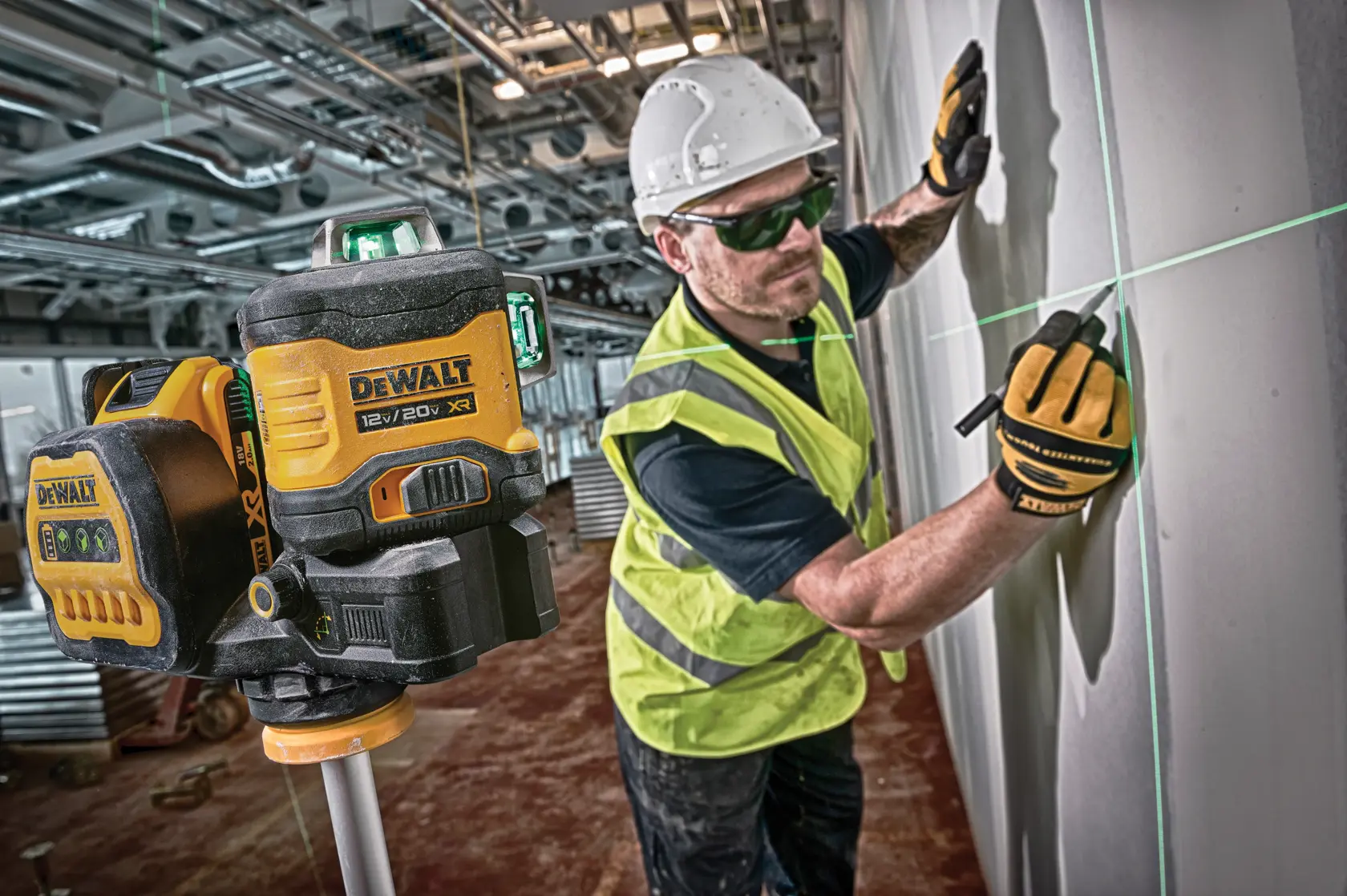 Un dispositivo de nivel láser DEWALT 12V/20V XR está colocado en un trípode en primer plano, proyectando líneas láser verdes sobre una pared. Al fondo, un trabajador de la construcción con equipo de seguridad, que incluye casco, guantes y chaleco de alta visibilidad, marca la pared utilizando las líneas láser como guía.