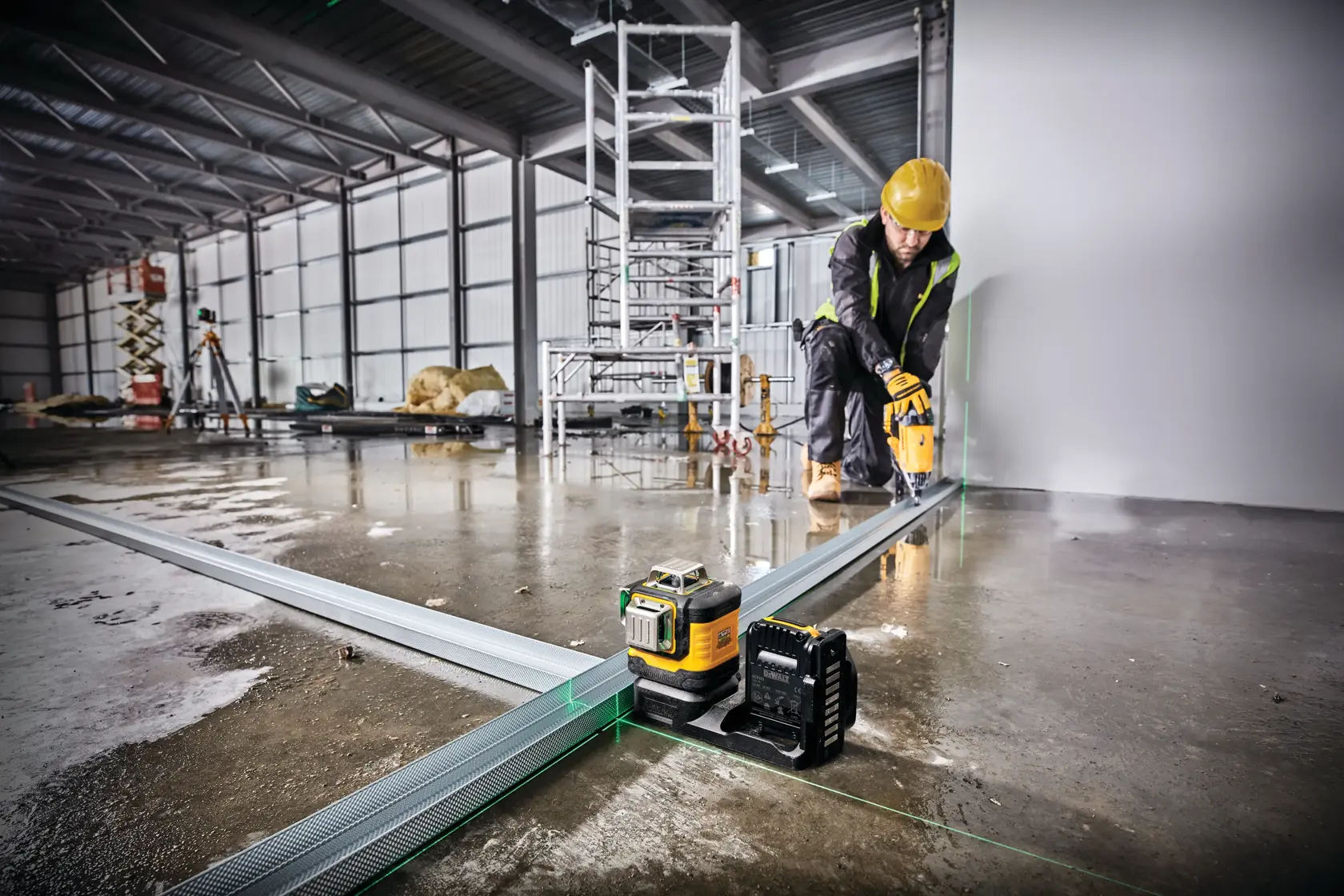 Una herramienta de nivel láser DEWALT está colocada en el suelo de una obra en construcción, proyectando líneas láser verdes a lo largo de rieles metálicos. Una persona con casco amarillo y equipo de seguridad trabaja al fondo cerca de la pared. En el sitio se ven andamios y materiales de construcción.