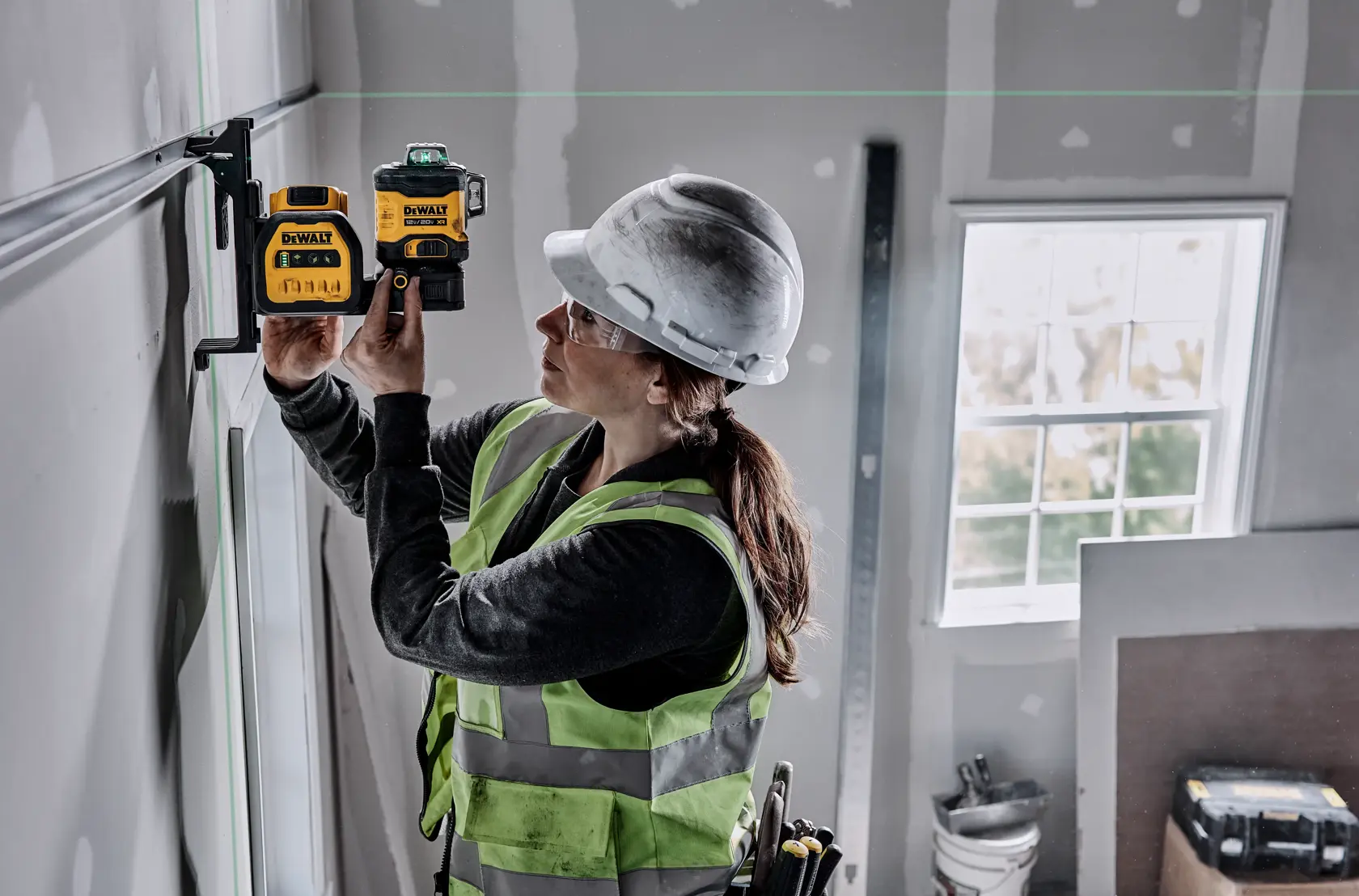 Una persona con casco y chaleco de seguridad utiliza un nivel láser de línea verde DEWALT (modelo DCLE34030G) para medir o alinear en una obra, fijándolo a un soporte de pared.
