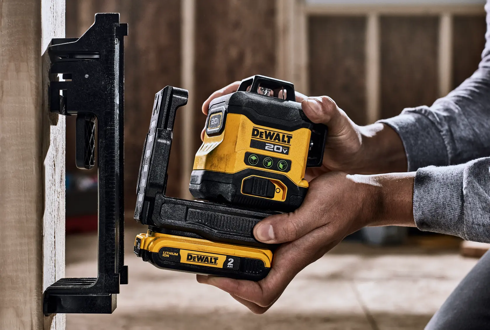 Una persona sostiene una herramienta de nivel láser DEWALT 20V amarilla y negra, utilizándola para alinearse con un soporte montado en la pared en un entorno de construcción.