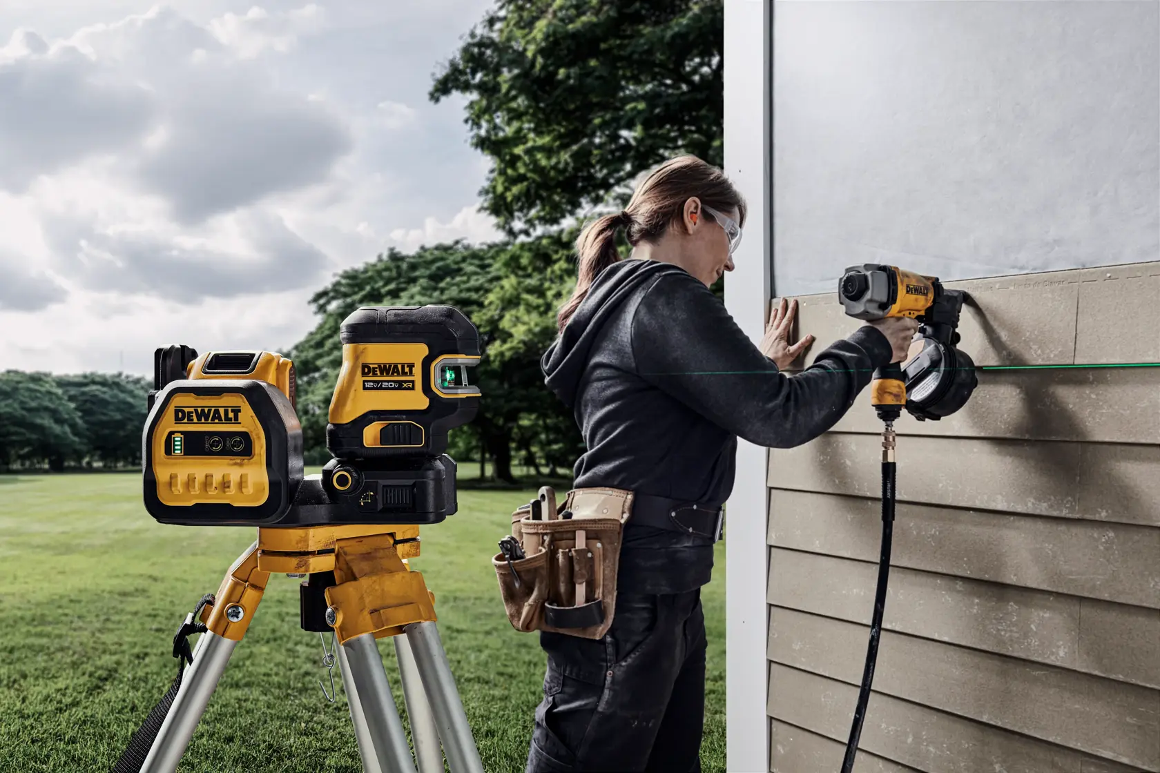 Una persona utiliza un nivel láser DEWALT y una herramienta eléctrica para instalar revestimiento en la pared exterior de un edificio, con el nivel láser DEWALT montado sobre un trípode en primer plano. La persona lleva un cinturón de herramientas y trabaja al aire libre.