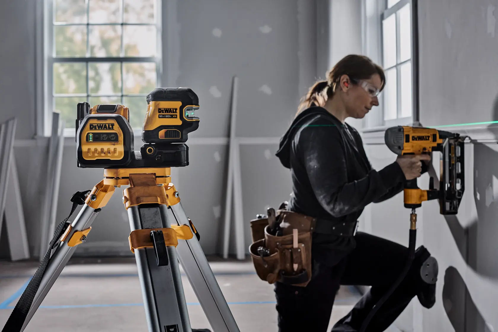 Una herramienta de nivel láser DEWALT está montada en un trípode en primer plano de una habitación en construcción. Al fondo, una persona con el rostro difuminado utiliza una herramienta eléctrica DEWALT para instalar material en una pared. Se pueden ver varios materiales y herramientas de construcción en la habitación.