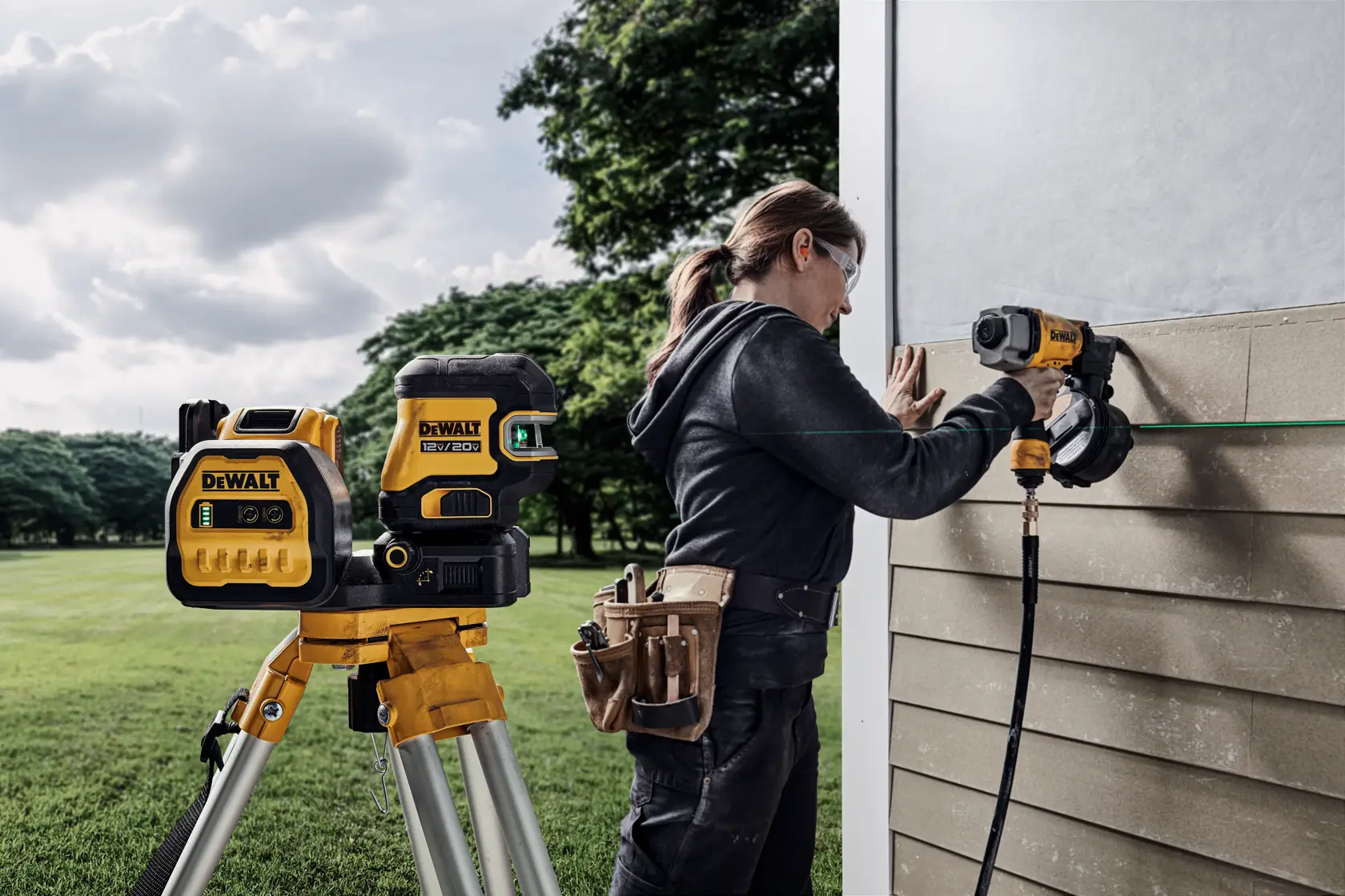 Una persona utiliza herramientas de construcción DEWALT, incluyendo un nivel láser montado en un trípode y una herramienta eléctrica, para trabajar en el lateral de un edificio al aire libre. La cara de la persona está desenfocada para preservar la privacidad, y lleva un cinturón de herramientas y ropa de trabajo. Se ven árboles y césped en el fondo.