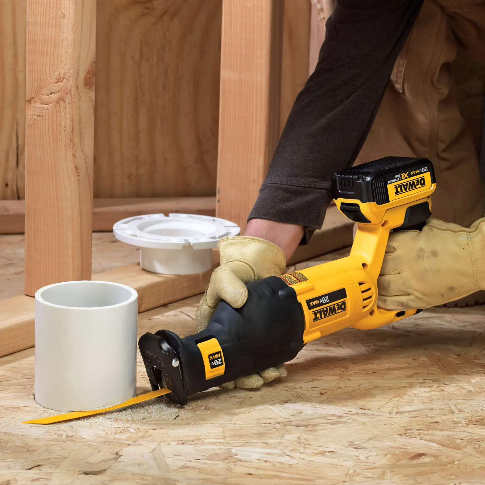 Una persona con guantes de trabajo utiliza una sierra recíproca inalámbrica DEWALT 20V Max para cortar un tubo de PVC blanco sobre un suelo de madera, con estructura de madera y componentes de fontanería visibles en el fondo.