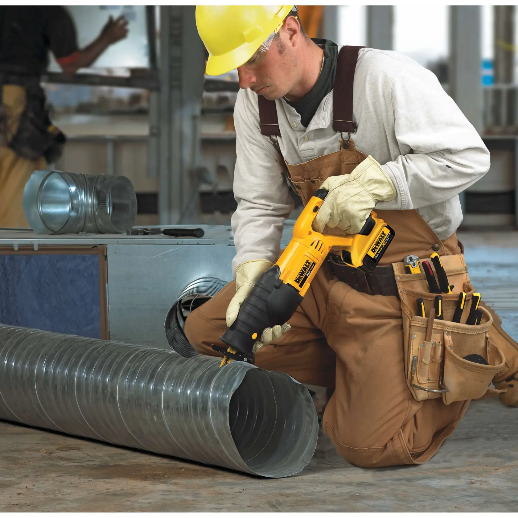 Un trabajador de la construcción con mono marrón, guantes y casco amarillo utiliza una sierra recíproca inalámbrica DEWALT (modelo DCS380B) para cortar un conducto metálico en un lugar de trabajo industrial. Lleva herramientas en una bolsa y está de rodillas rodeado de componentes metálicos.